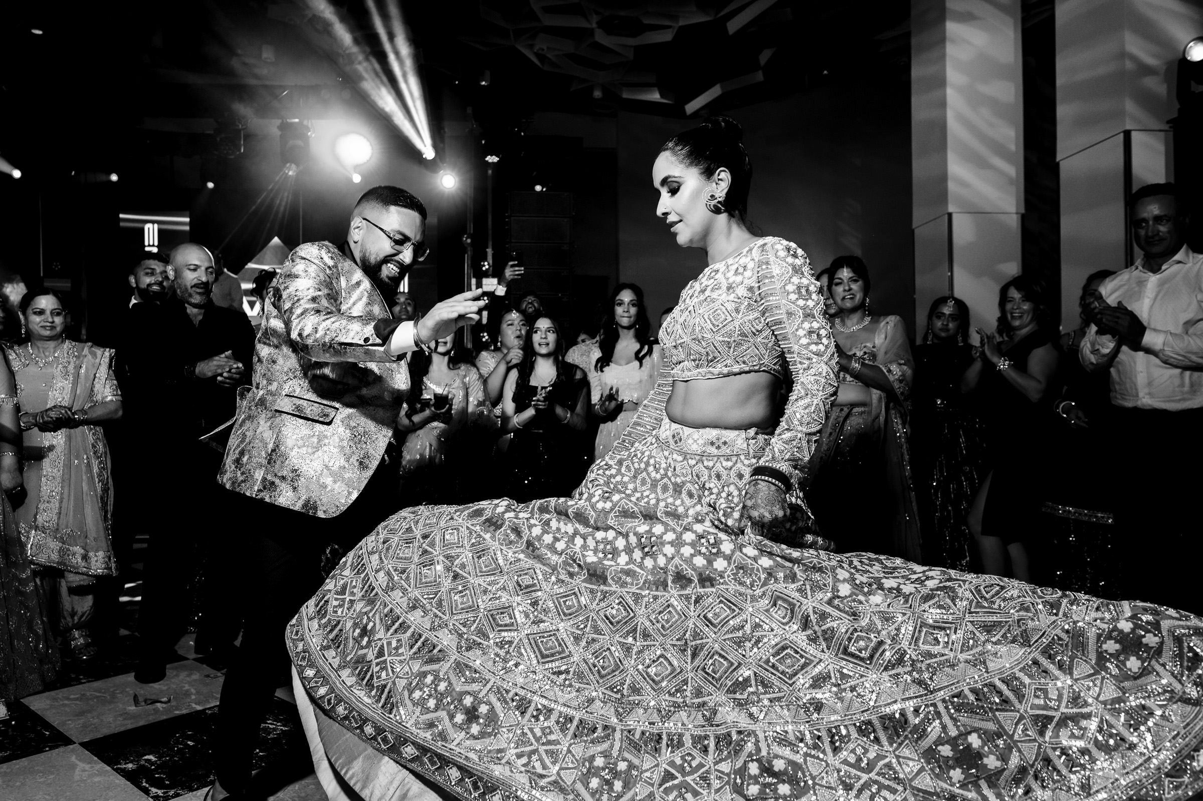 A man in a shiny blazer dances energetically with a woman in an intricate, flowing lehenga, reminiscent of favourite wedding photos. Surrounded by a cheering crowd at this lively party, the black and white scene beautifully captures the joy and movement.
