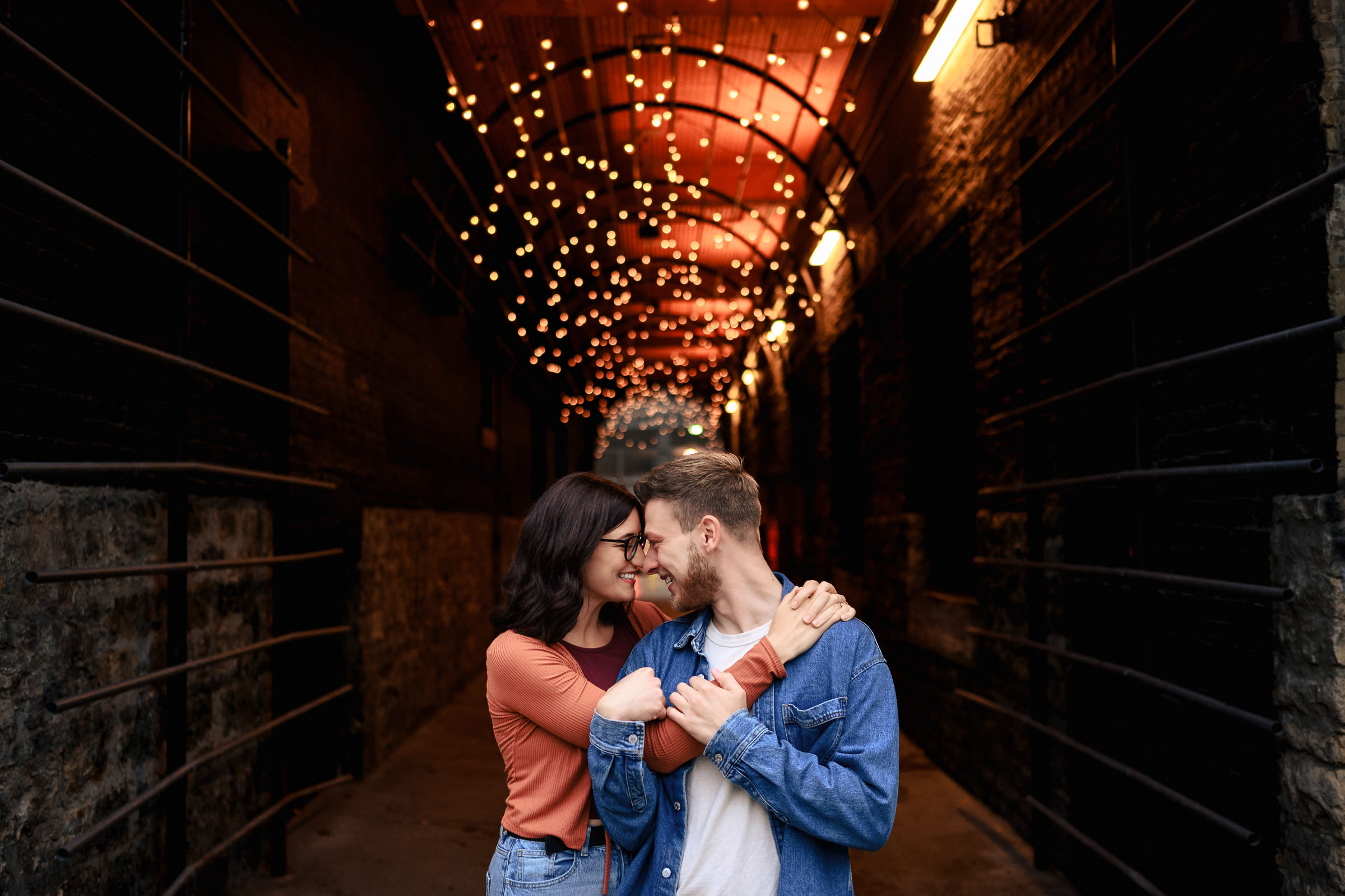 A couple embraces in a dimly lit alleyway adorned with string lights, capturing one of their favorite wedding photos. The man in a denim jacket and the woman in an orange top stand against stone walls, creating a cozy and romantic atmosphere.