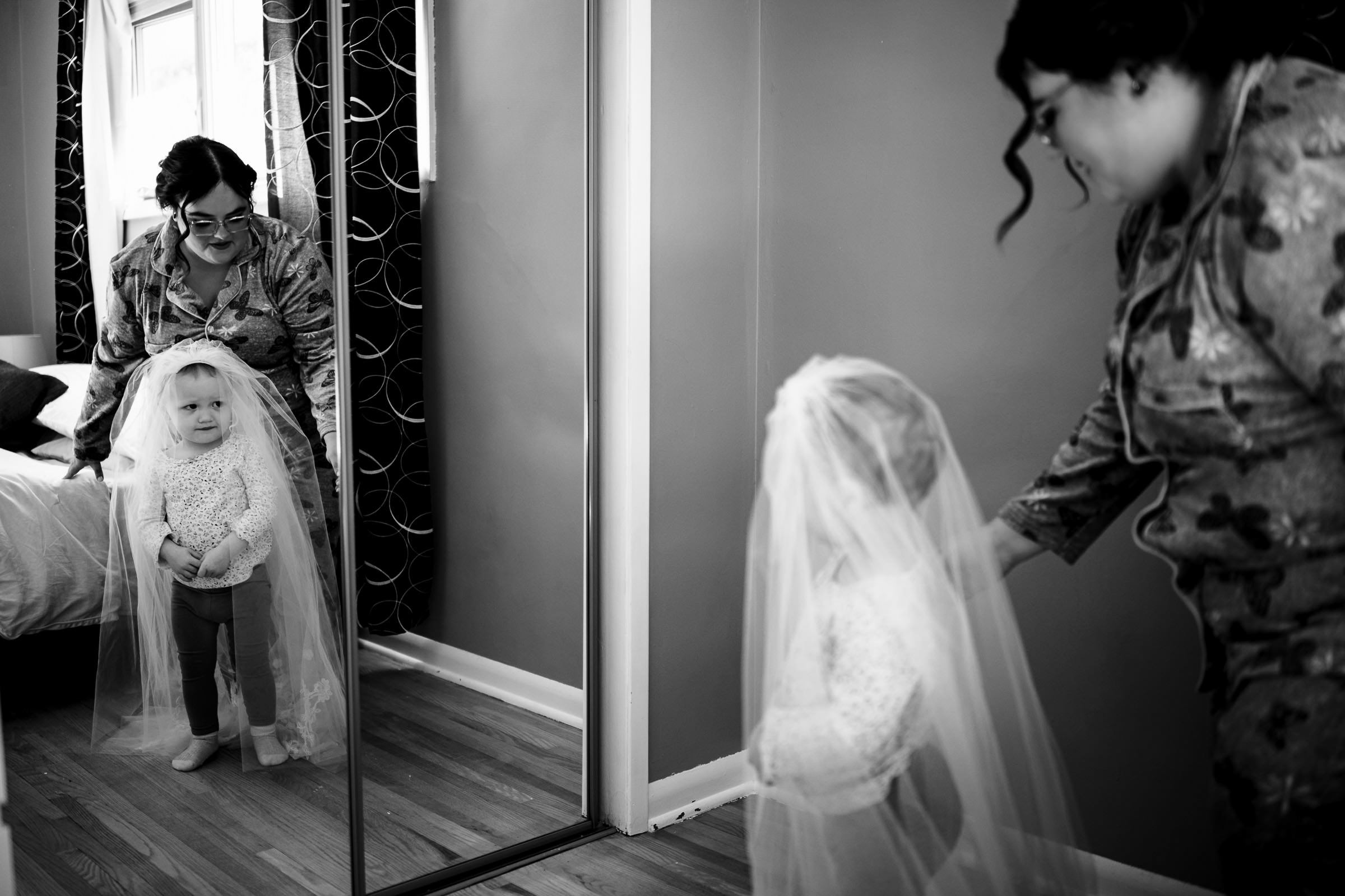 A young child stands in front of a mirror, wearing a bridal veil, lost in their reflection. An adult nearby, dressed casually, gently adjusts the veil. The scene, reminiscent of favourite wedding photos, unfolds on a wooden floor with a patterned curtain backdrop. Black and white photo.