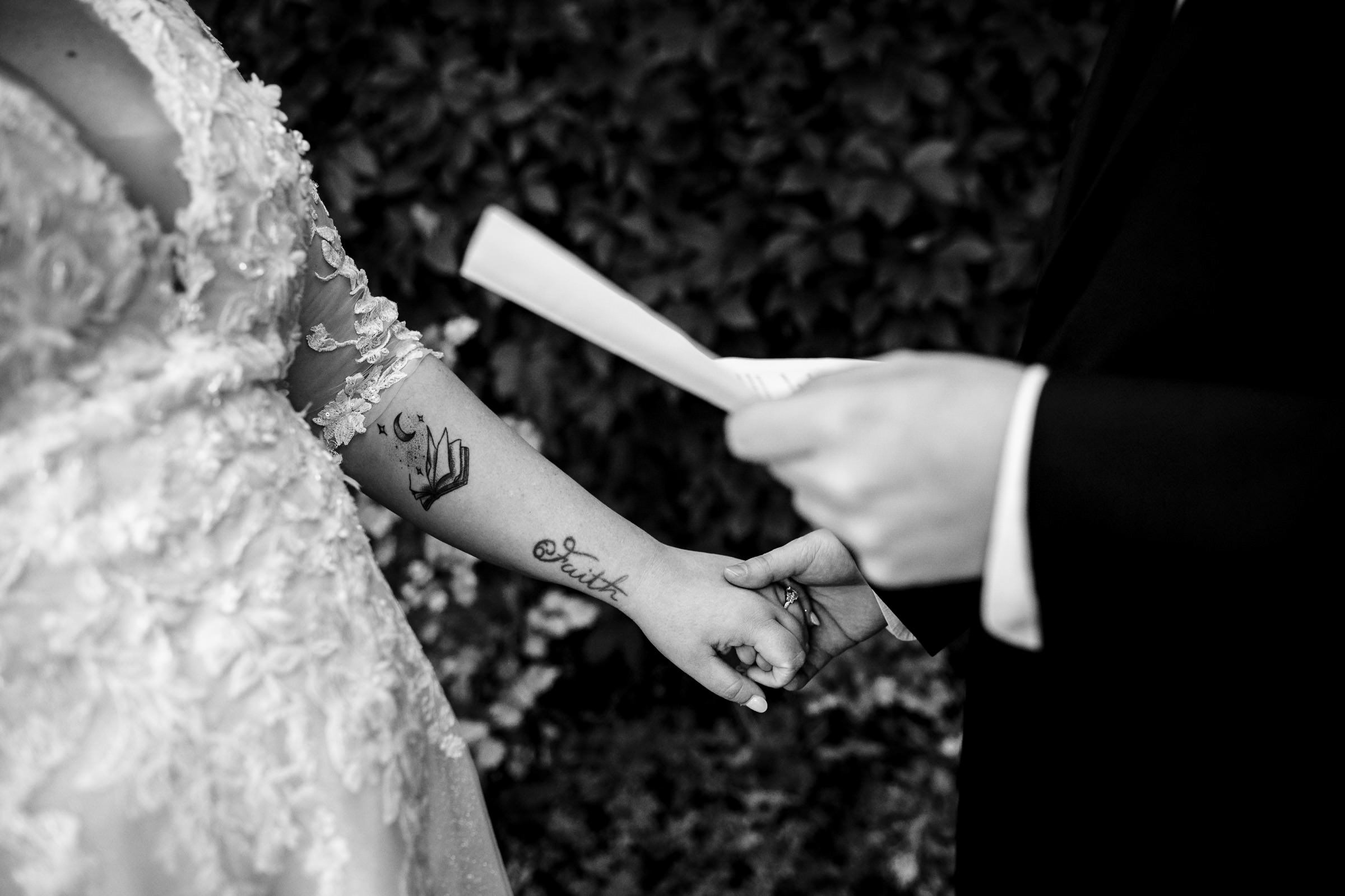 A bride and groom hold hands during an outdoor ceremony, captured in one of their favourite wedding photos. The bride's lacy dress reveals a tattoo on her forearm, while the groom clutches a piece of paper. Foliage adds depth to the black and white image.