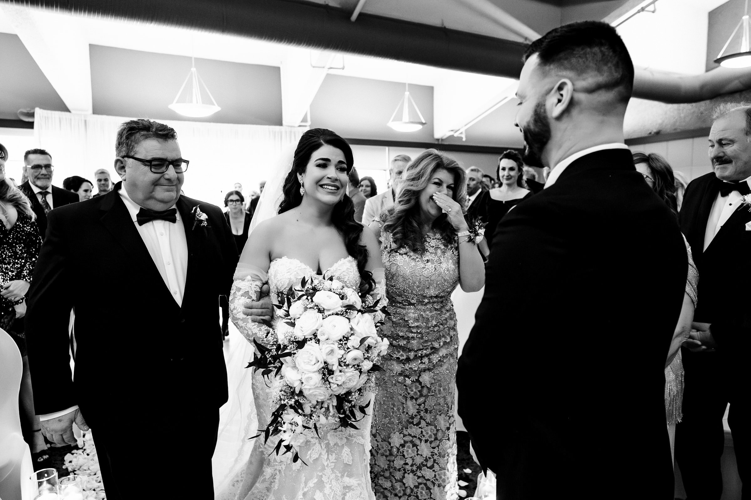 A bride holding a bouquet walks down the aisle with a man and a woman, who appears emotional—a scene sure to be among the favourite wedding photos. A groom stands at the altar, smiling. Guests are seated in the background. The setting is indoors with bright lighting and large fixtures.