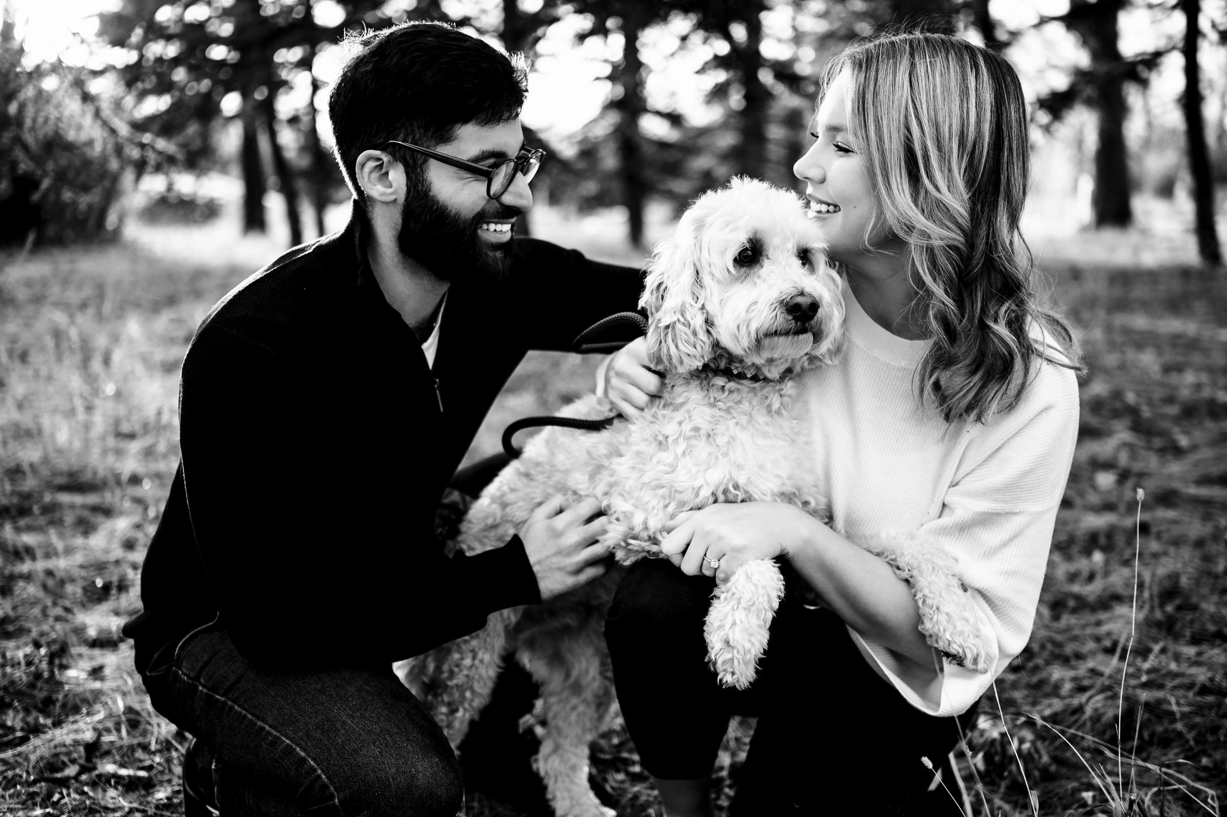 A man and a woman smile at each other while holding a fluffy dog between them, standing outdoors in a wooded area with trees in the background. This black-and-white image captures one of their favourite wedding photos, embodying joy and togetherness.