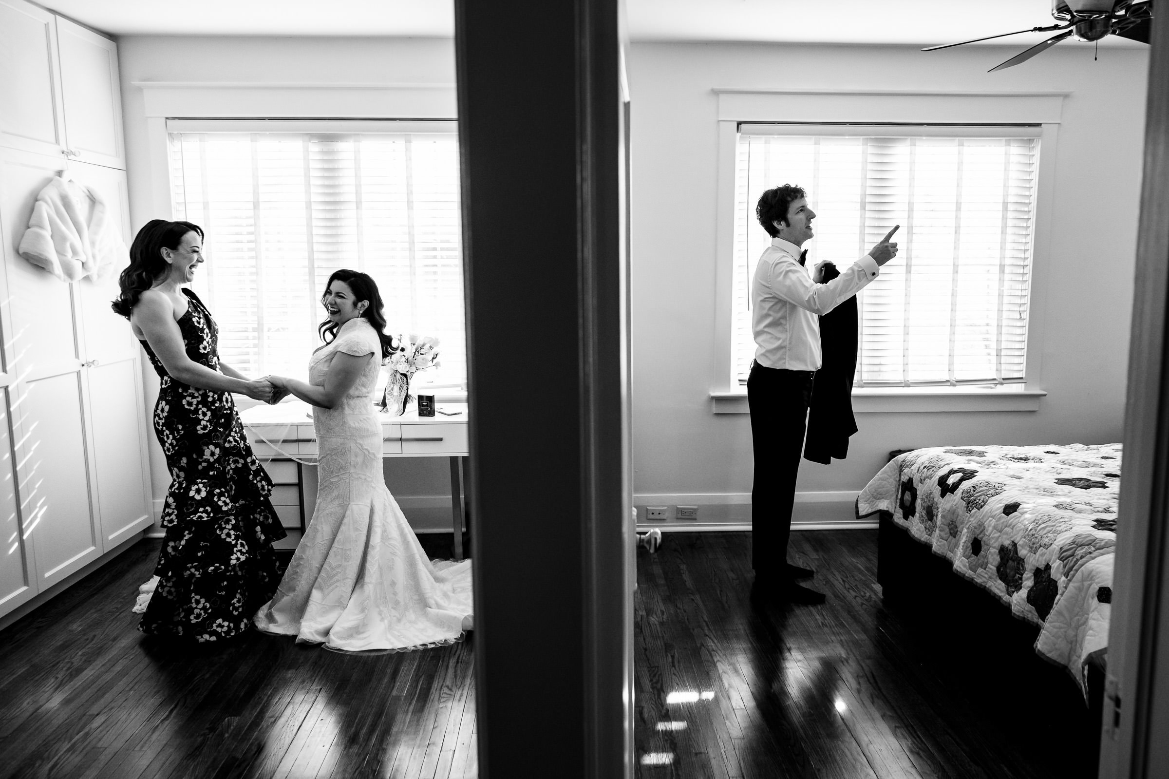 A black and white photo captures one of my favourite wedding moments: a bride in a stunning dress stands beside a woman in florals on the left side of a divided room. On the right, a man readies himself, jacket in hand, as sunlight pours through large windows into both bright rooms.