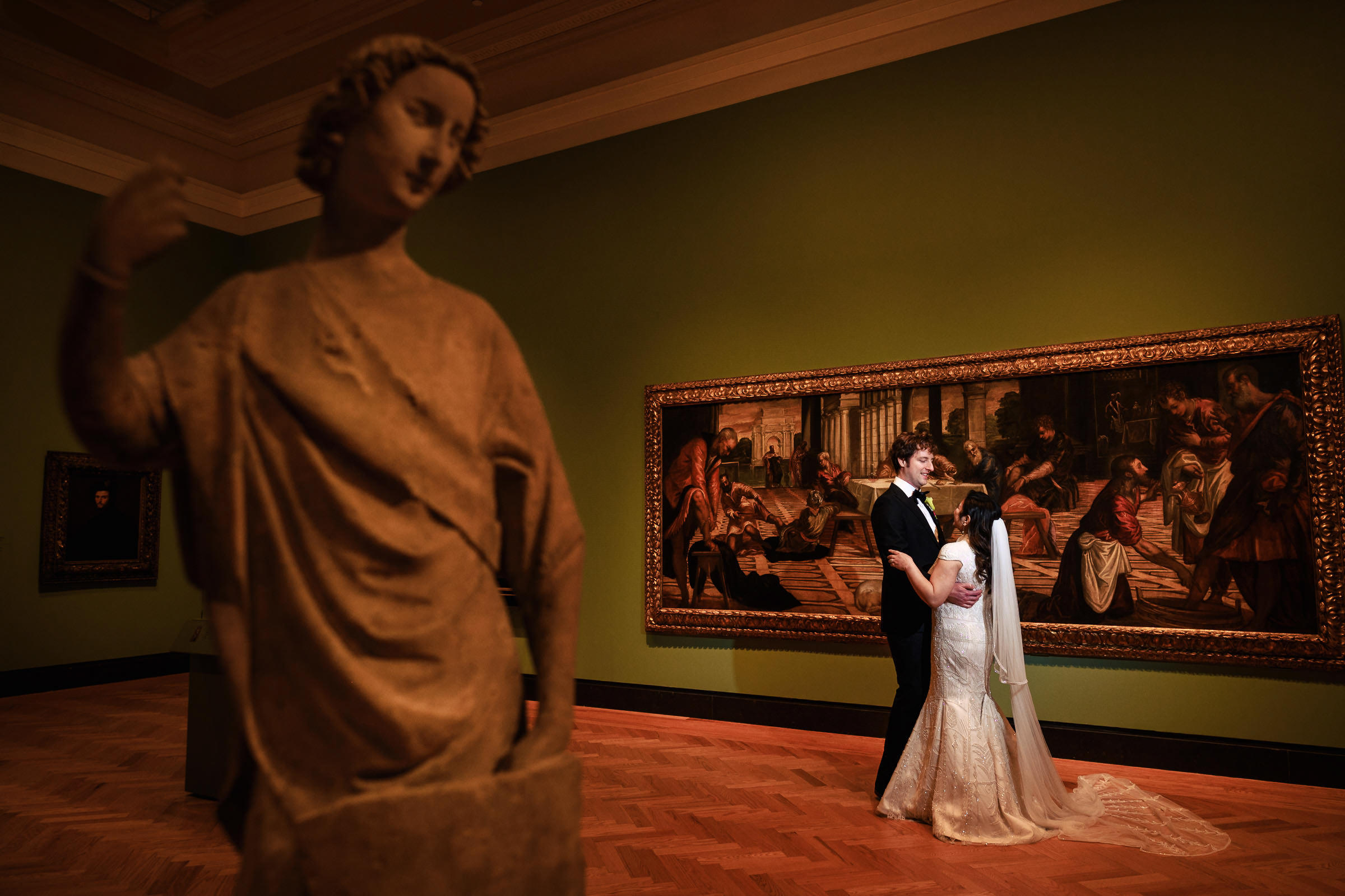 A bride and groom share a tender moment, embracing in a gallery with green walls—a scene destined to become one of their favourite wedding photos. A classical statue graces the foreground on the left, while a large framed painting adorns the wall behind them.