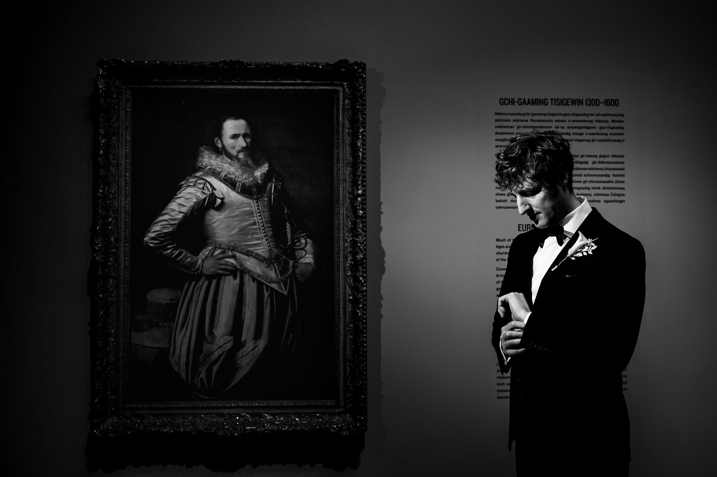 A man in formal attire stands in a dimly lit room, gazing at an item in his hands. Beside him is a framed painting of a historical figure in elaborate clothing. The setting exudes a classic, contemplative atmosphere, reminiscent of browsing through favourite wedding photos from the past.
