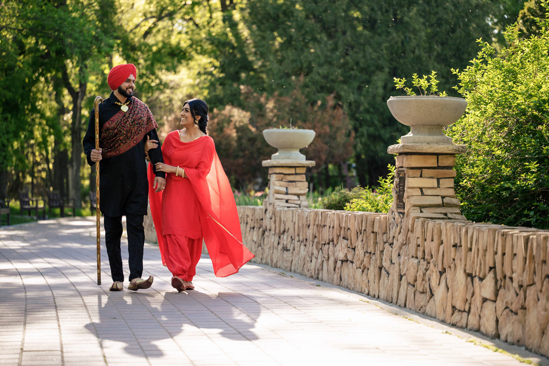 A couple in traditional attire walks along a sunlit path in a garden.