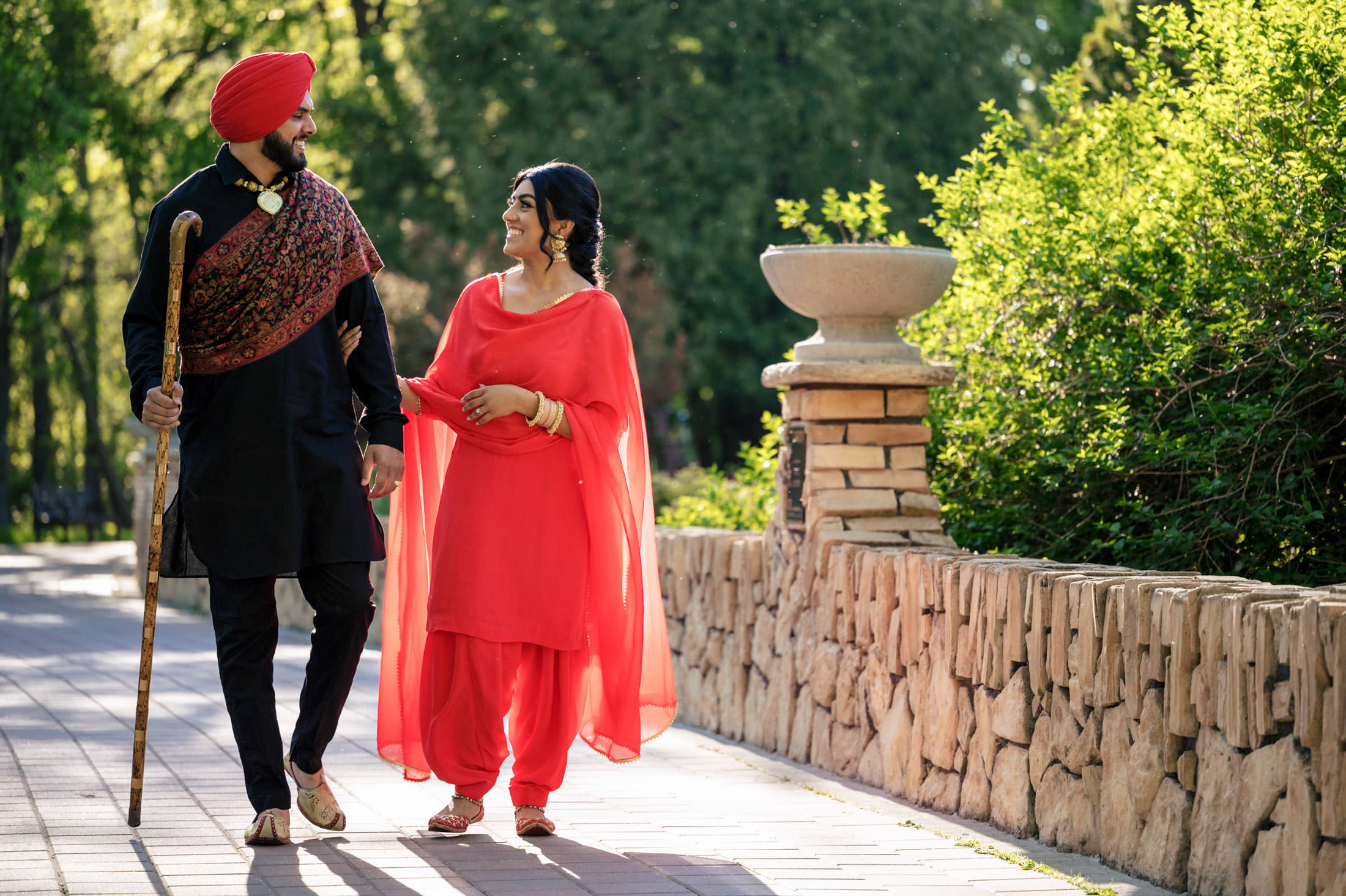 A couple in traditional attire strolls in a sunlit park, surrounded by greenery.