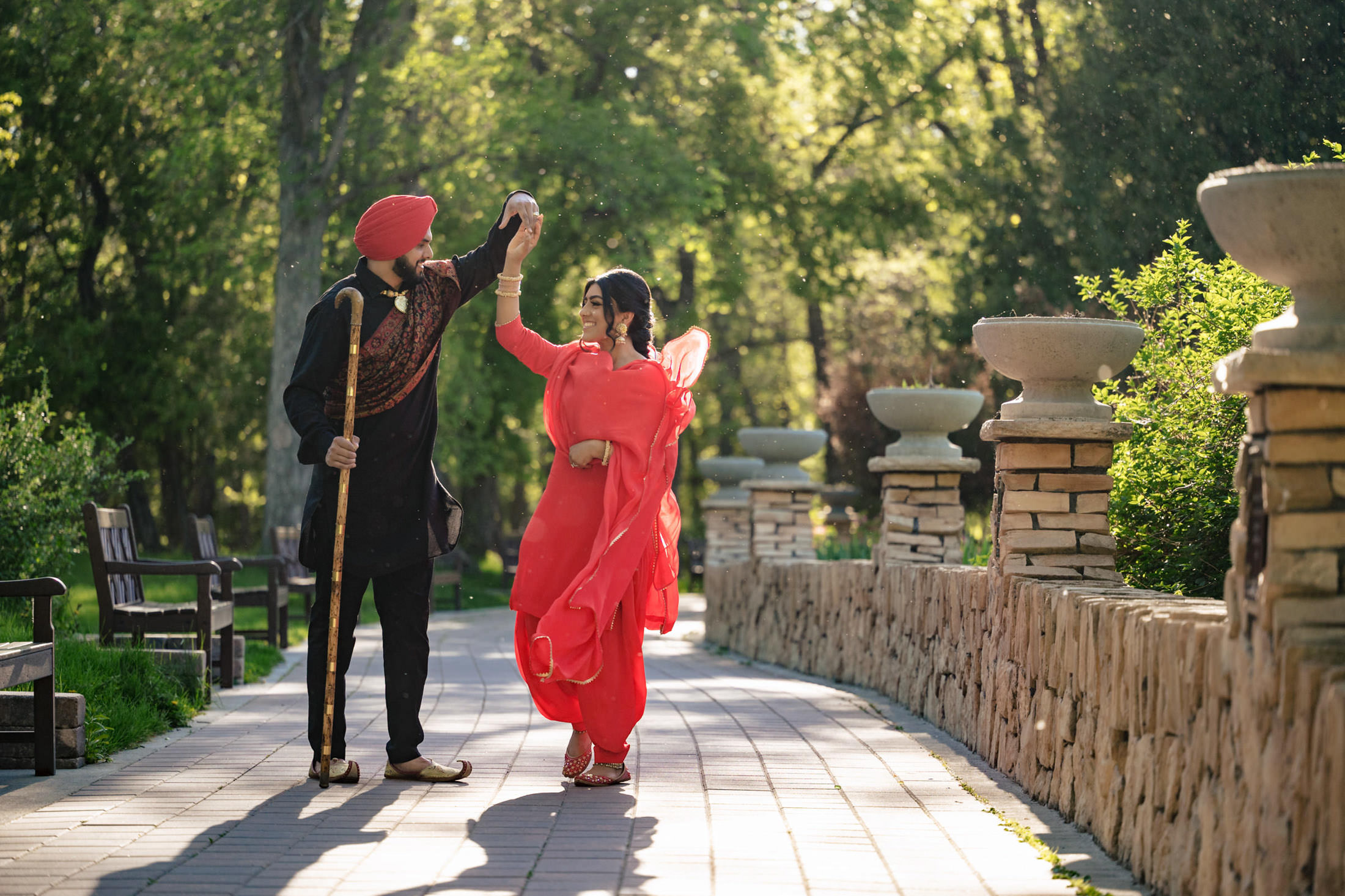 Couple dancing outdoors; man in turban, woman in red dress, sunny day, lush green background.