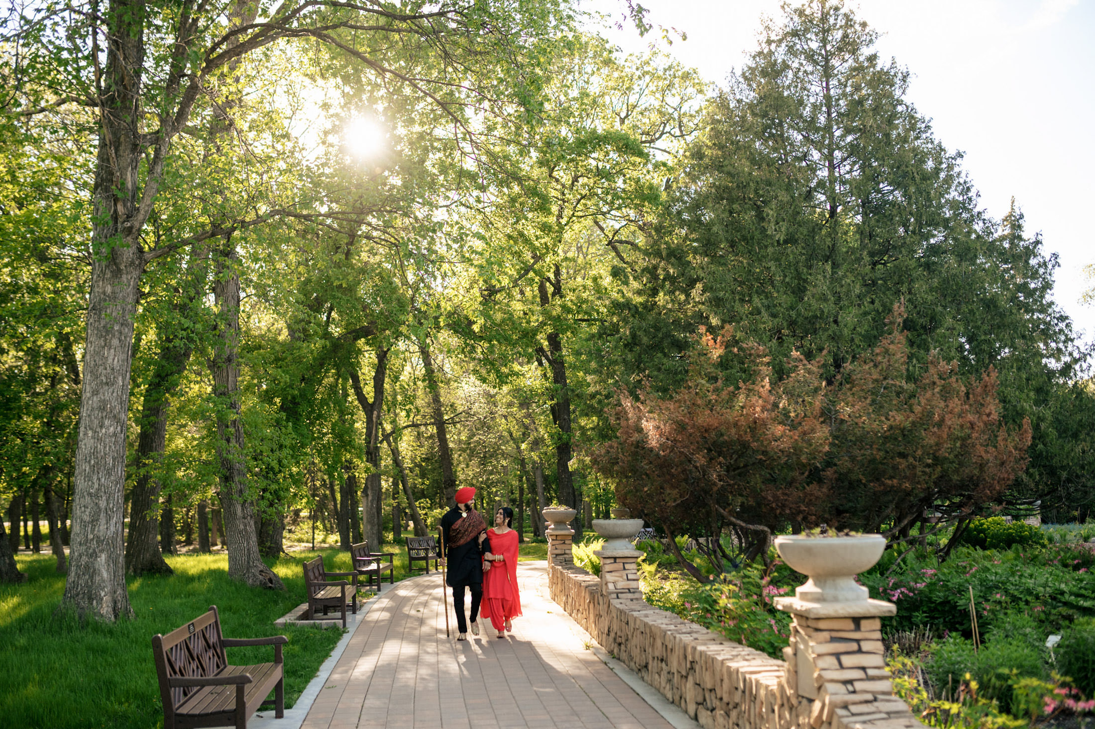 A couple walks hand in hand down a sunlit, tree-lined park path.