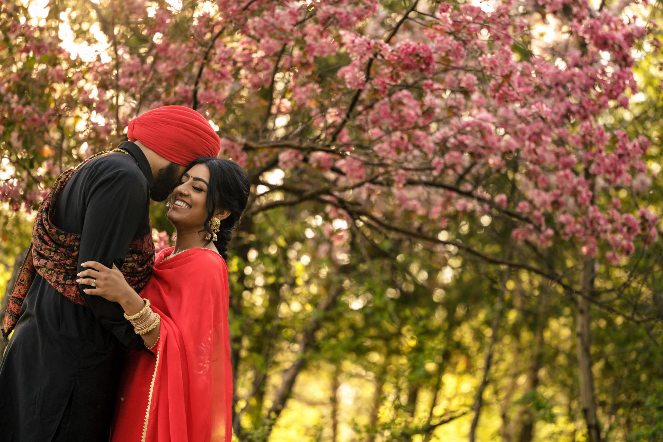 Couple in traditional attire embrace under blooming cherry blossoms in a sunlit garden.