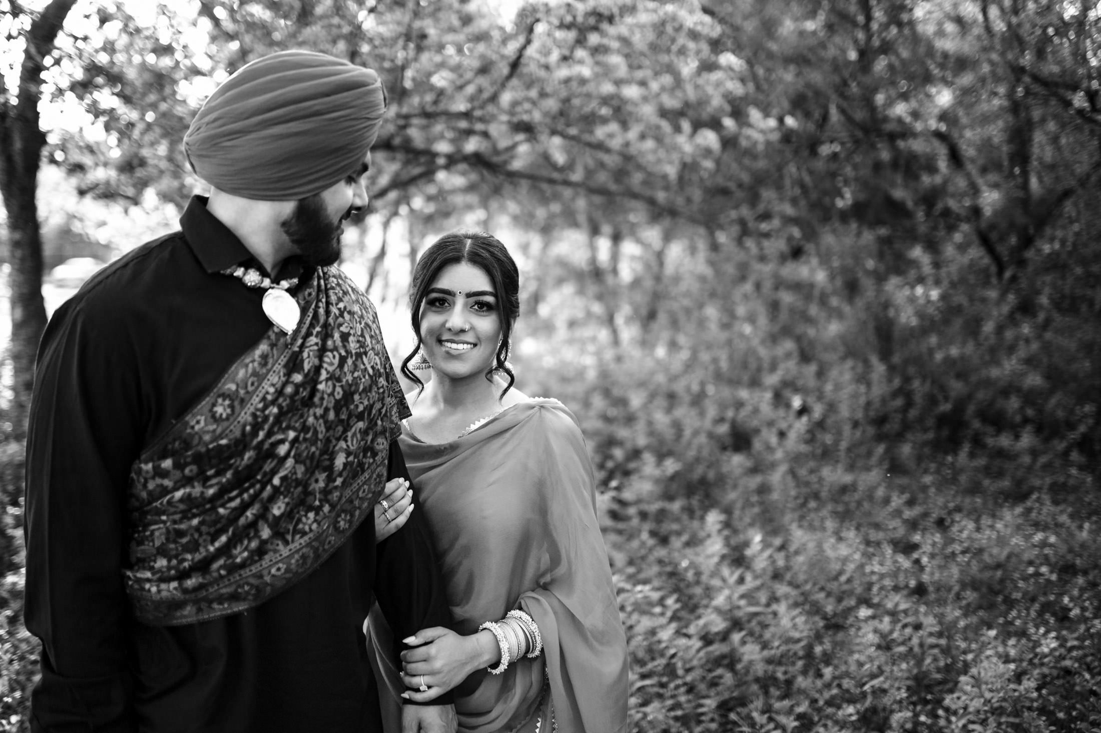 A couple in traditional attire smiles in a forested area, with trees and plants in the background.
