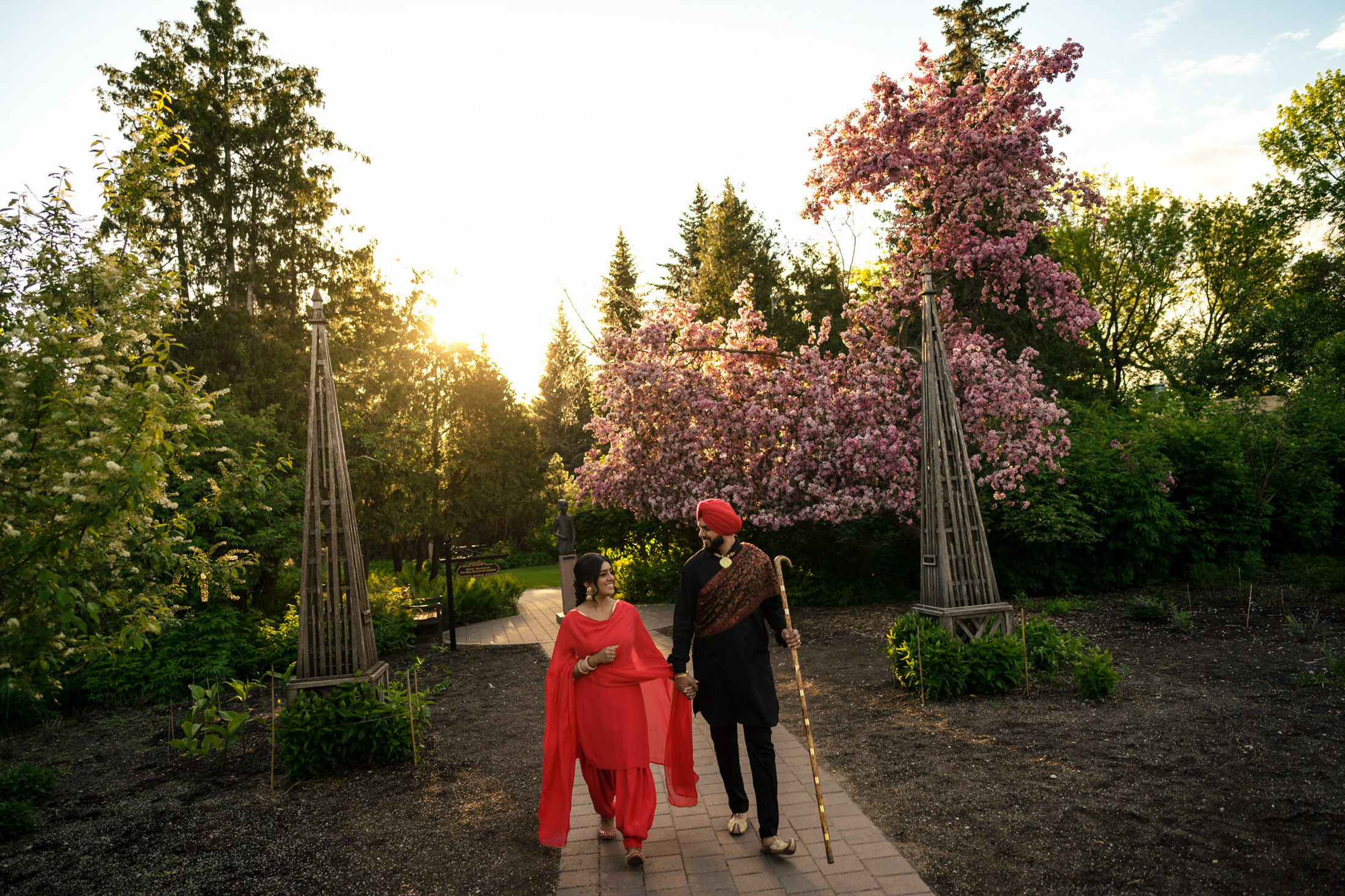 A couple in traditional attire walking through a garden with blooming trees at sunset.