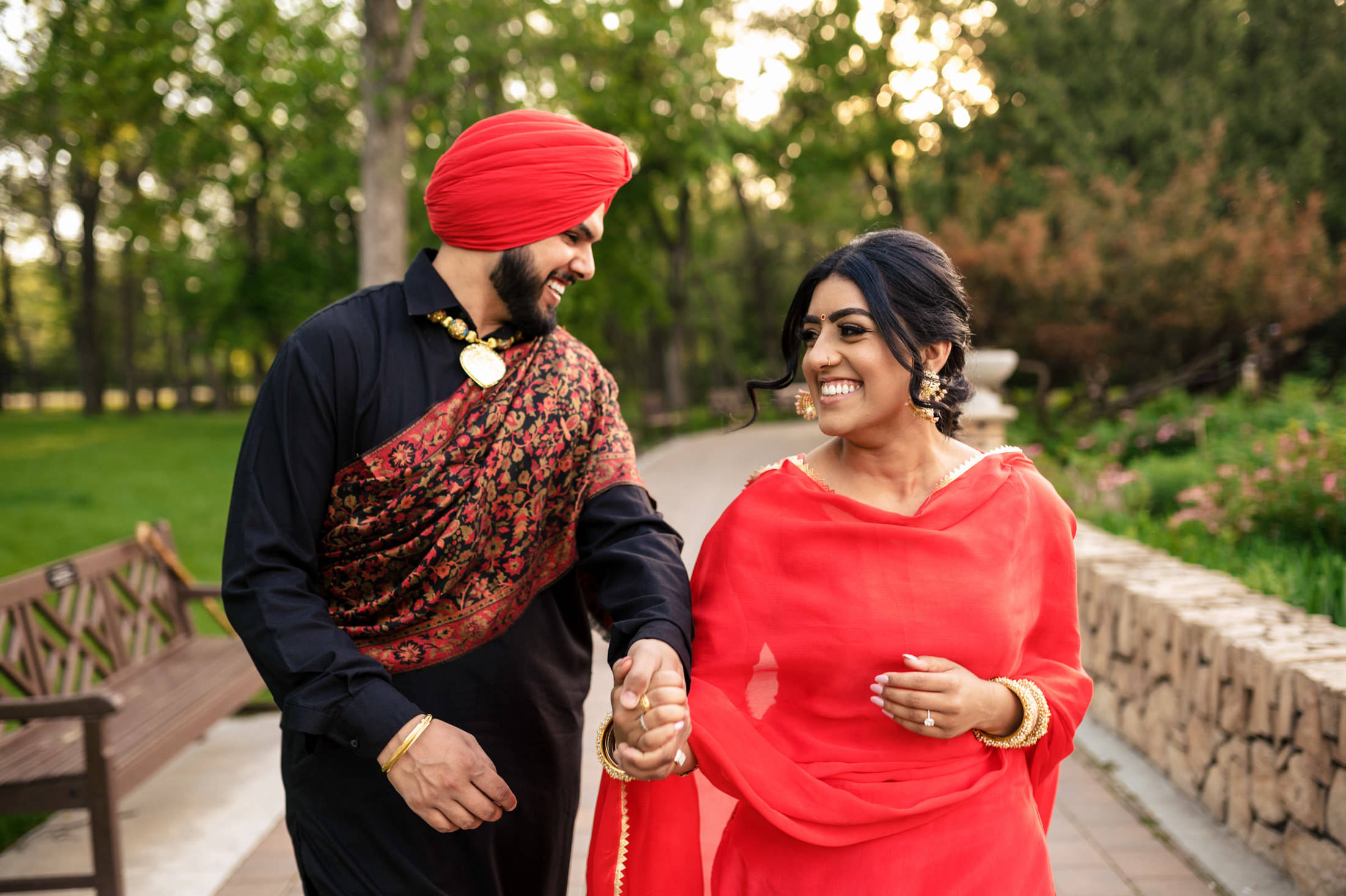 A couple in traditional red and black attire walking and smiling in a park setting.
