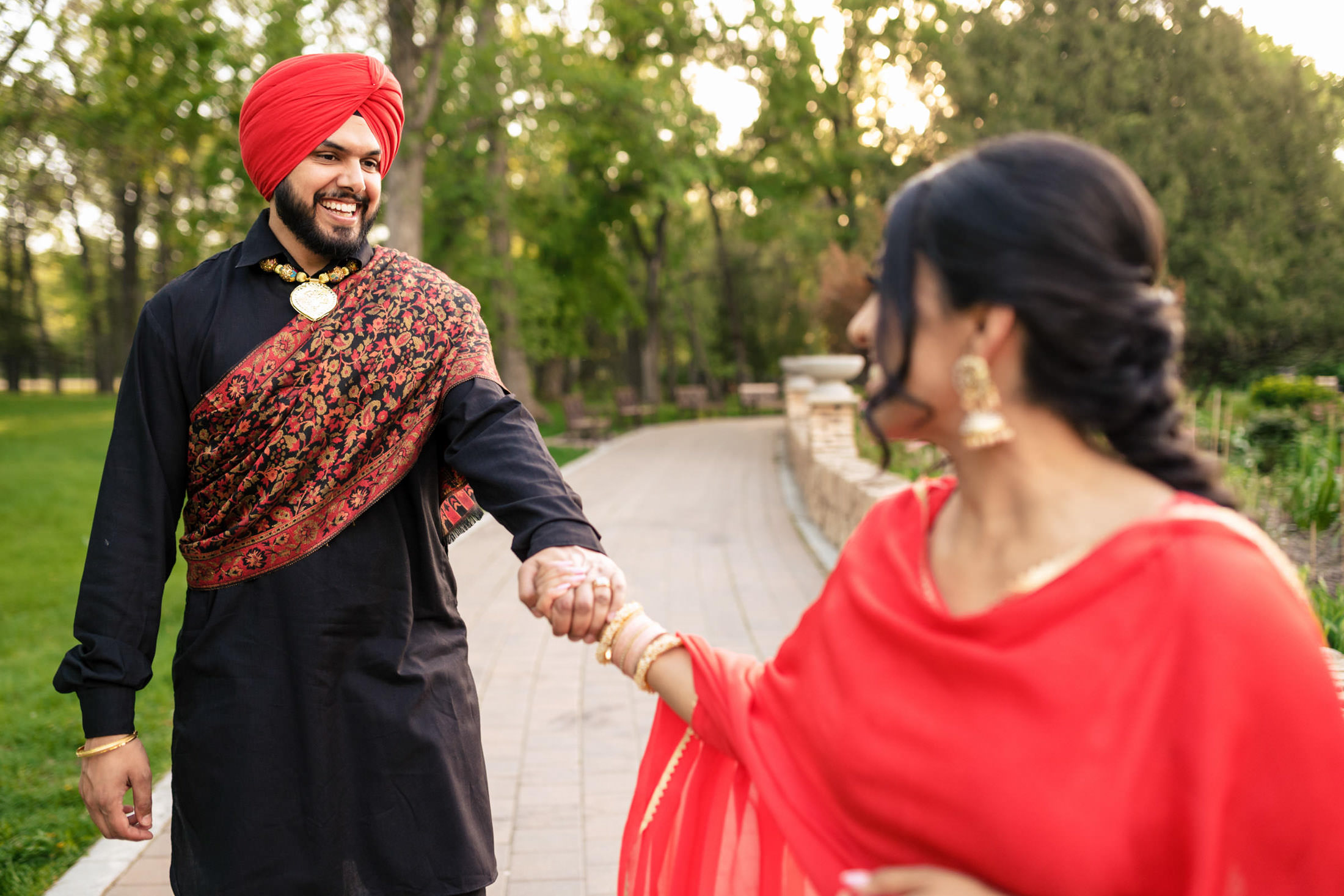 Man in red turban and woman in red dress hold hands on a park path.