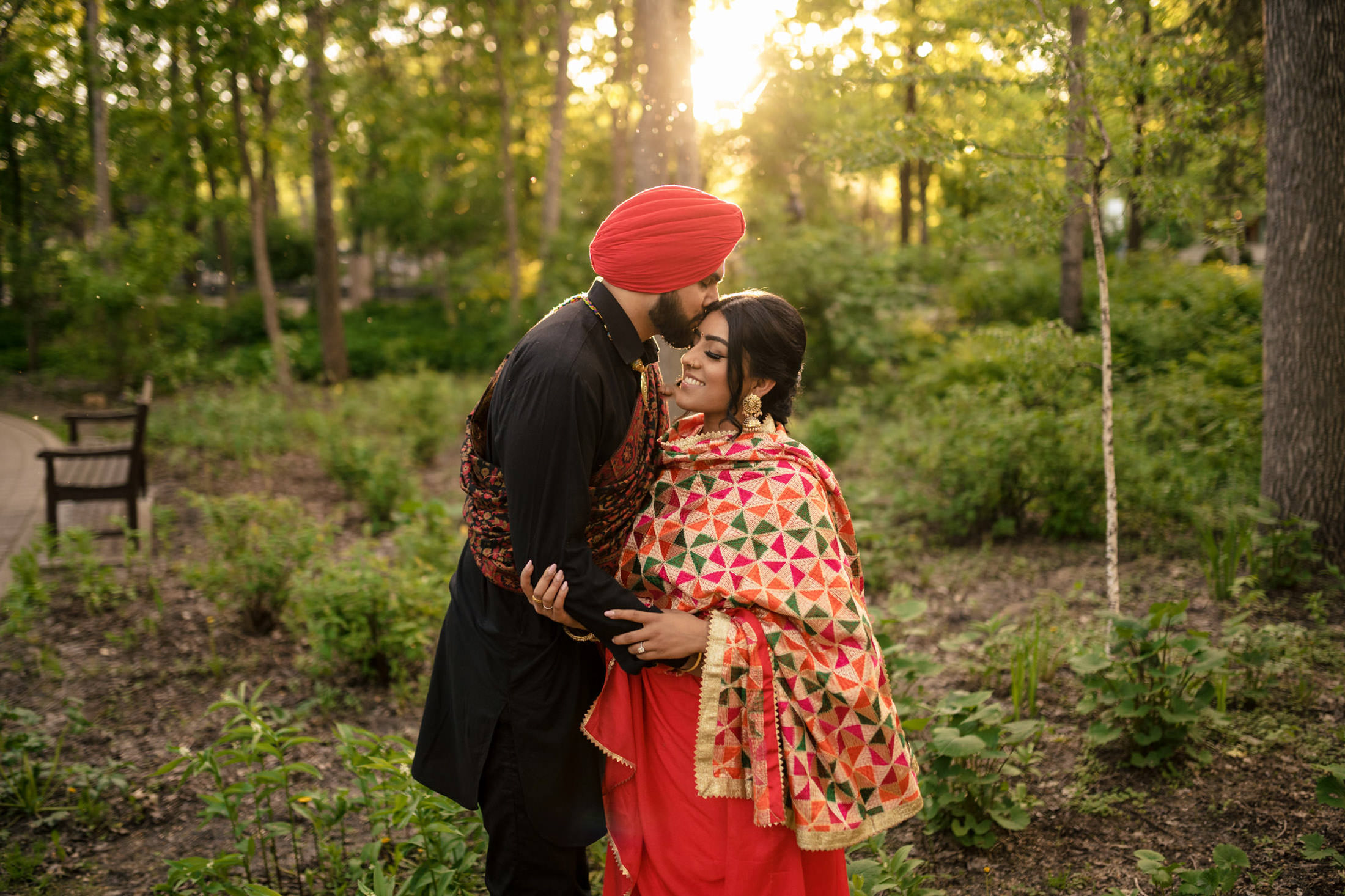 Couple in traditional attire embracing in a sunlit forest setting.