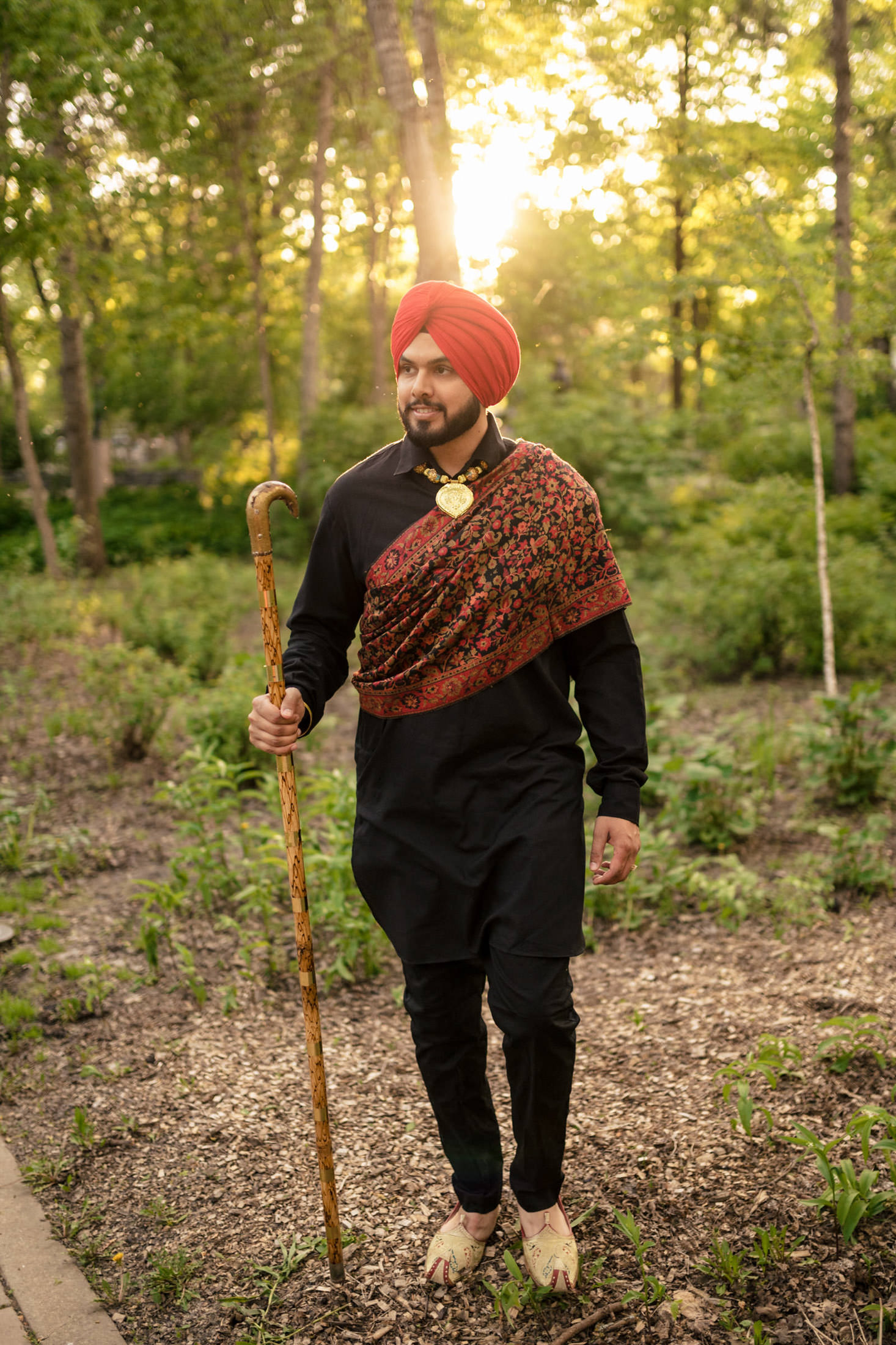 Man in red turban with a staff, dressed in traditional attire, walks in a sunlit forest.