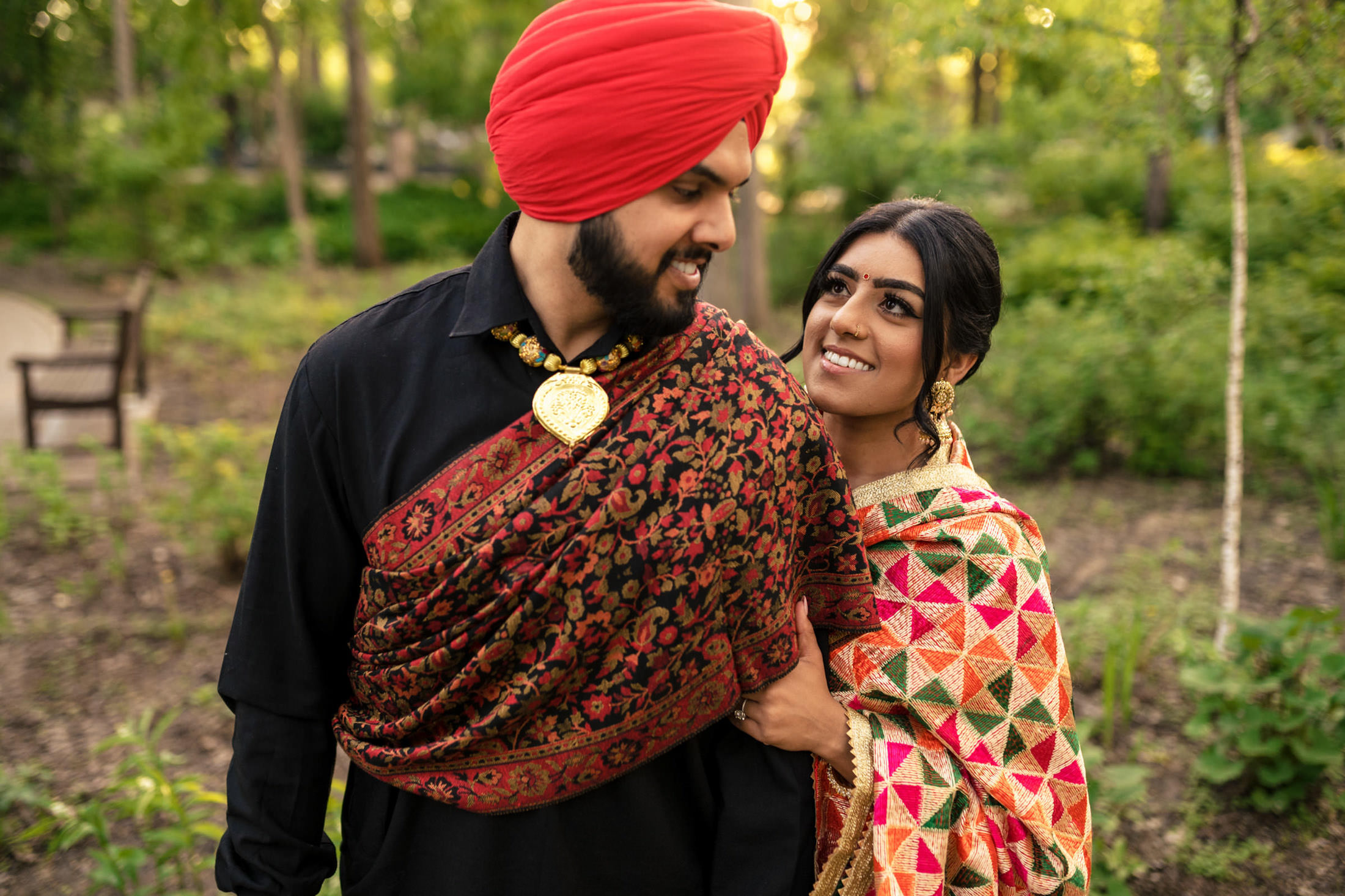A couple in traditional attire lovingly gaze at each other in a lush outdoor setting.