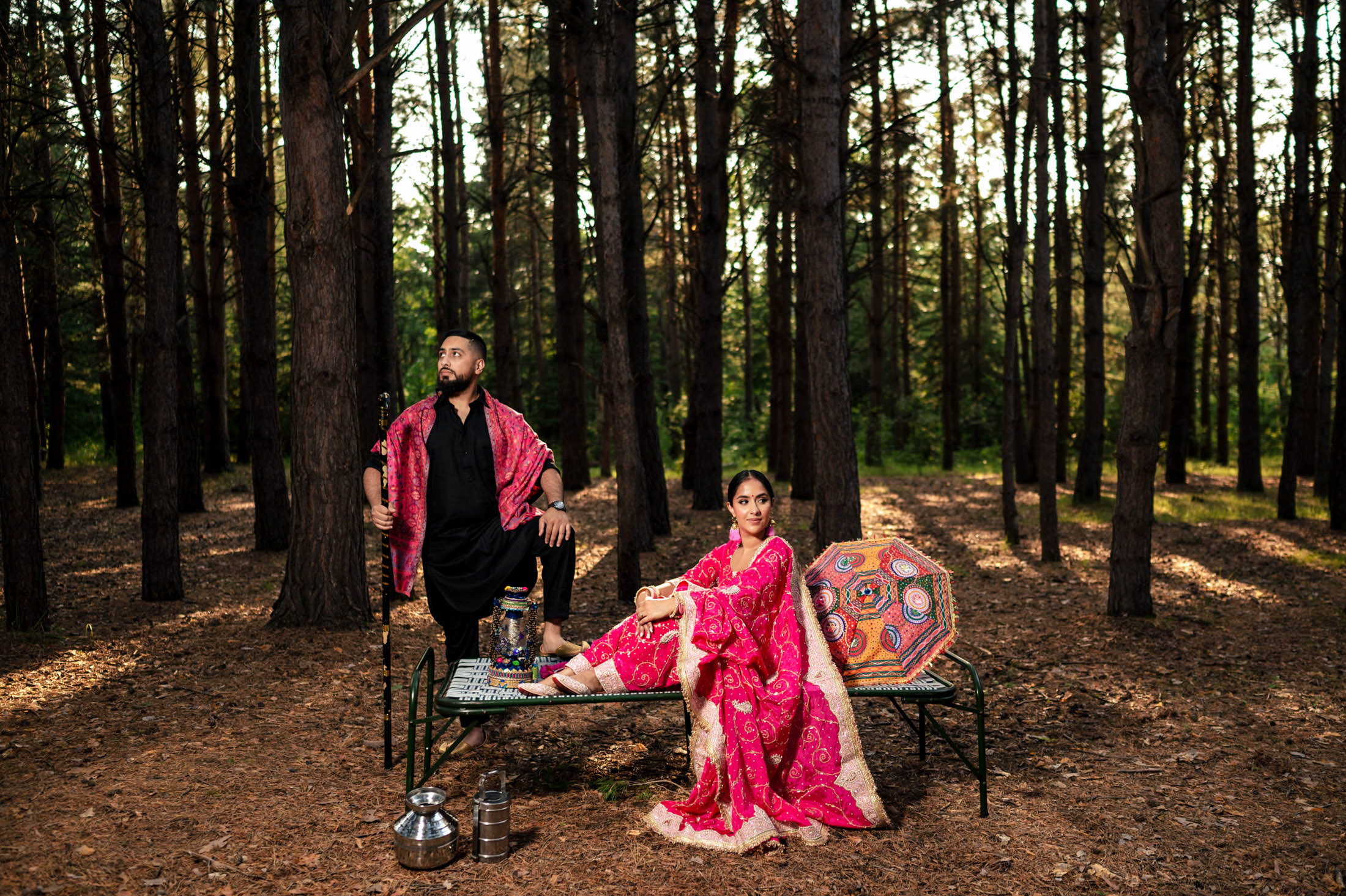 A couple in traditional attire poses elegantly on a bench in a forest.