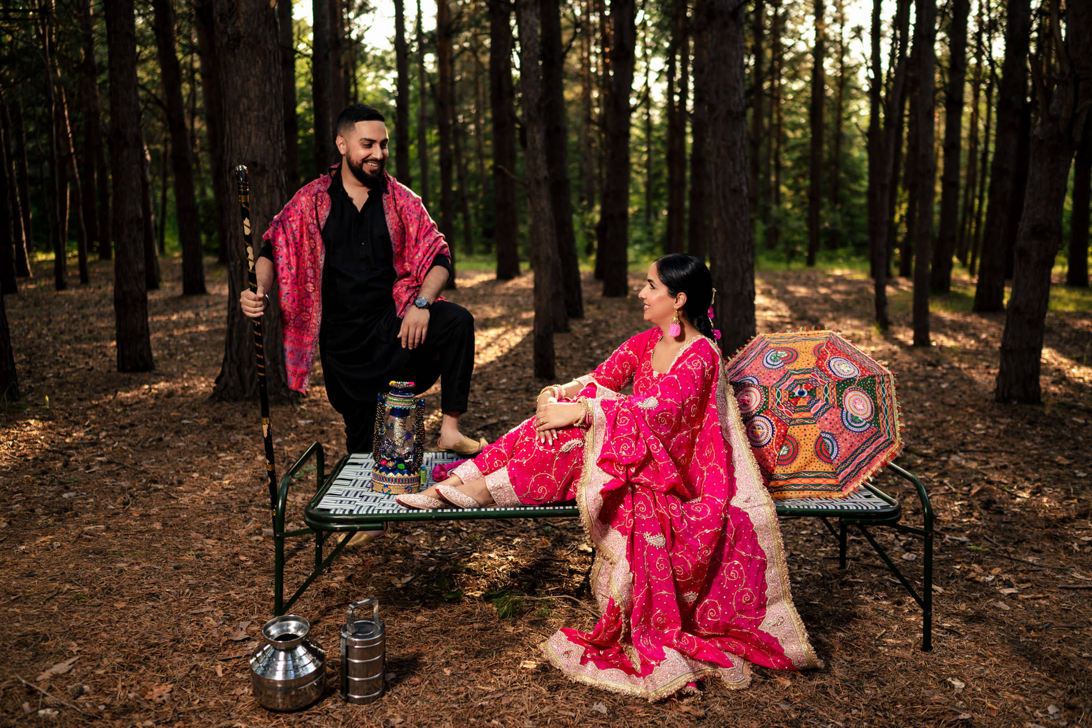A man and woman in traditional attire sit on a bench in a forest.