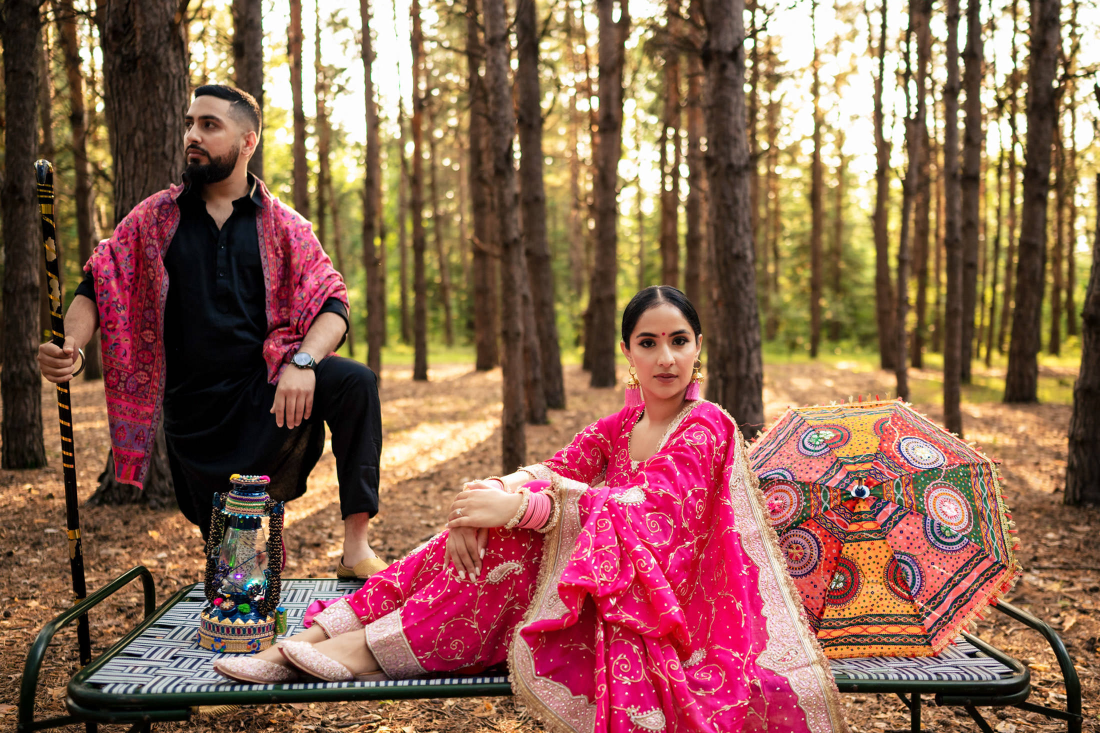 A man and woman in vibrant attire pose in a sunlit forest setting.