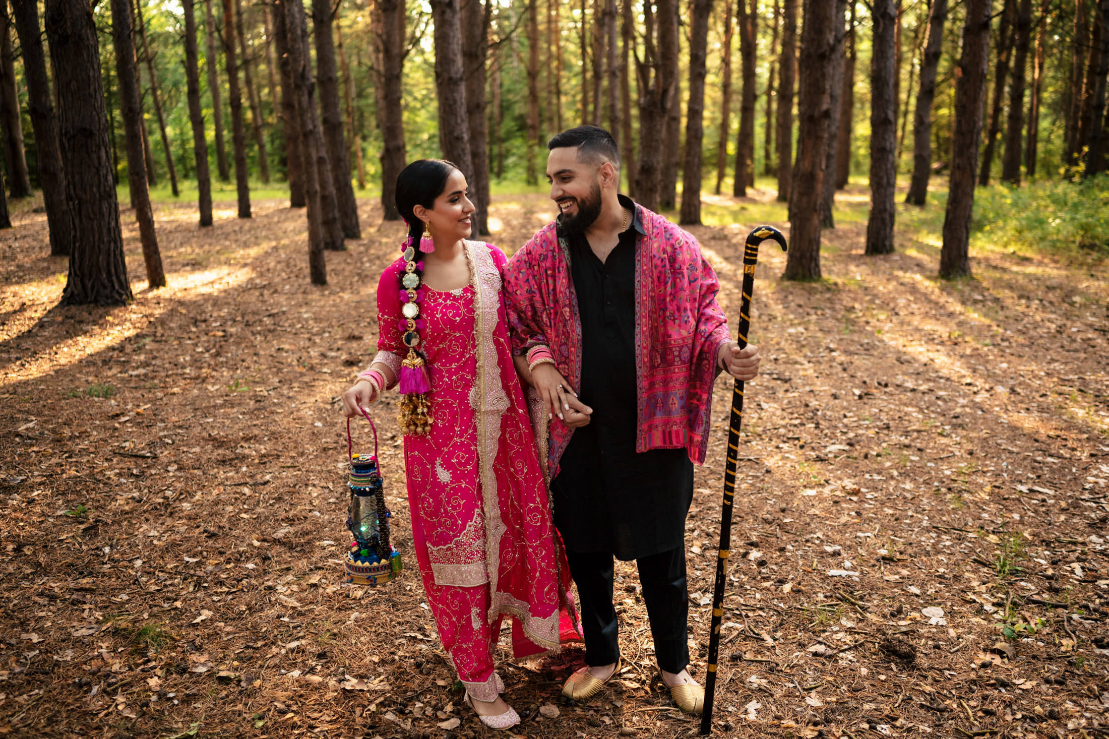 Couple in colorful attire walking in a sunlit forest, holding a lantern and a staff.