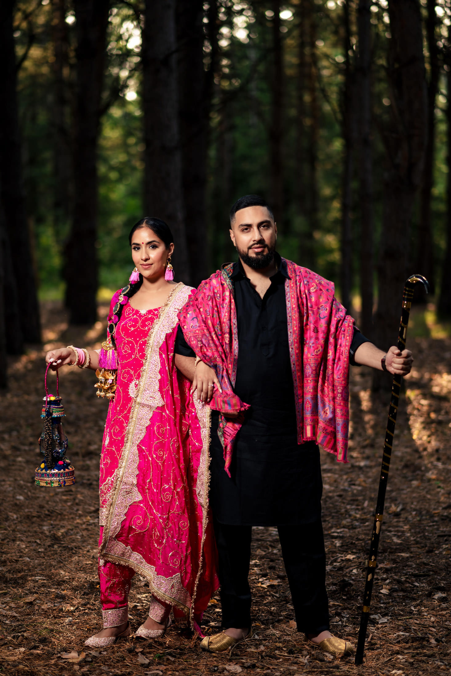 Couple in vibrant attire stands in a wooded area, holding a staff and decorative tassels.