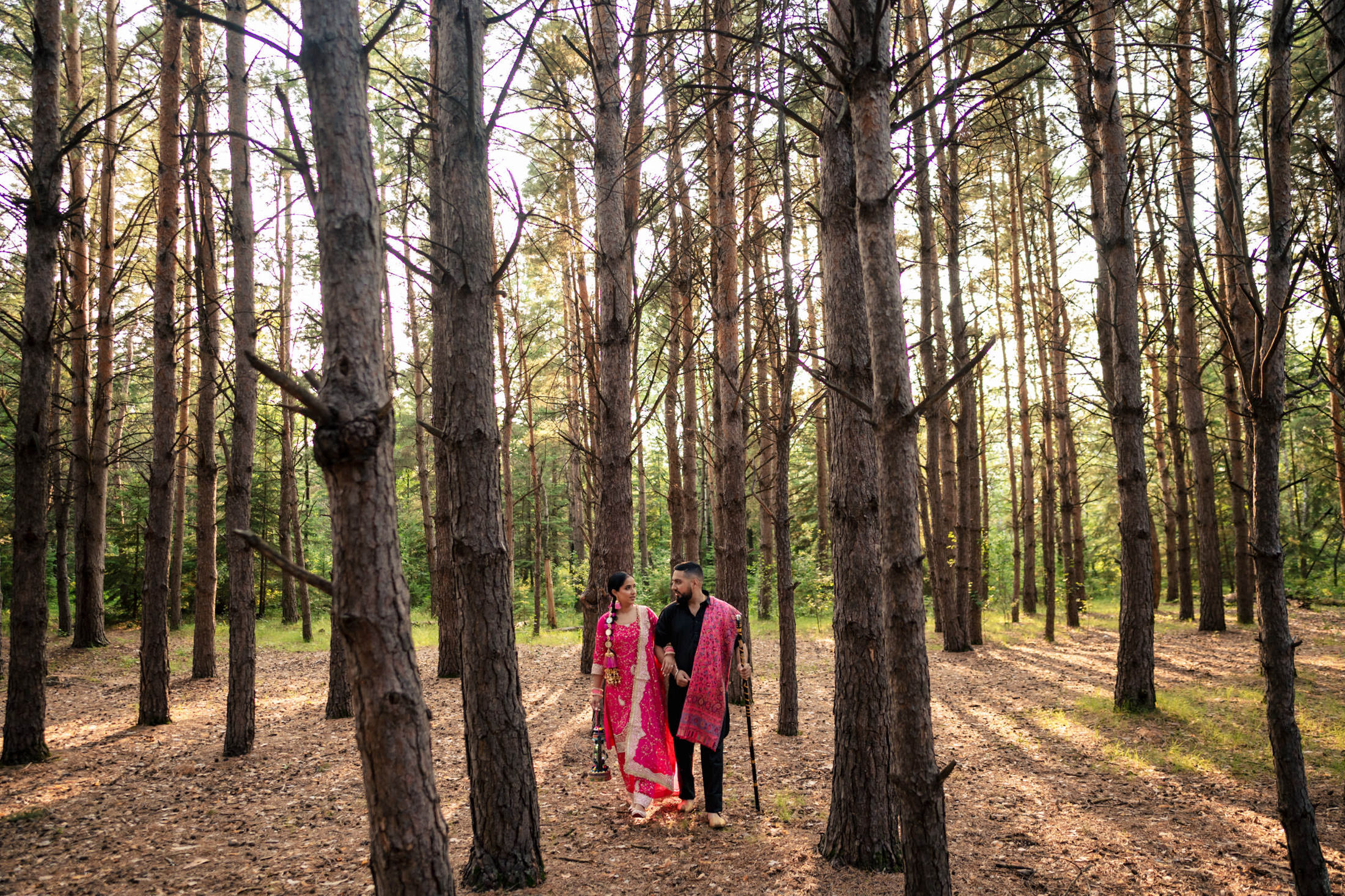 Couple in traditional attire walking through a sunlit forest with tall trees.