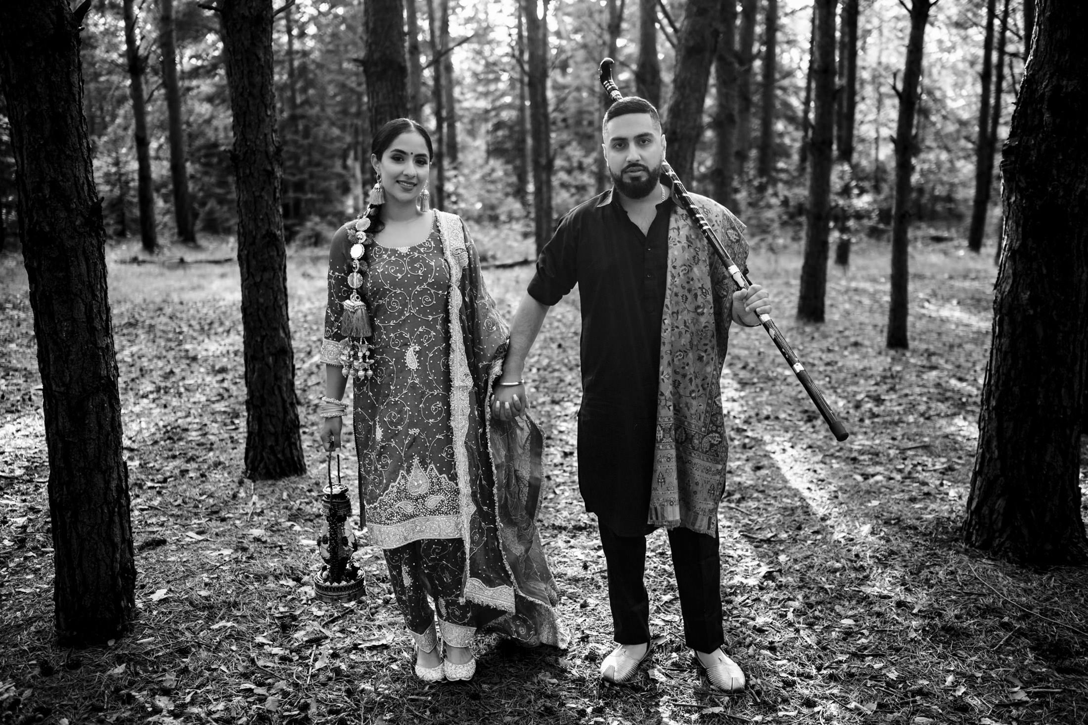 A couple in traditional attire stands in a wooded area, holding hands.