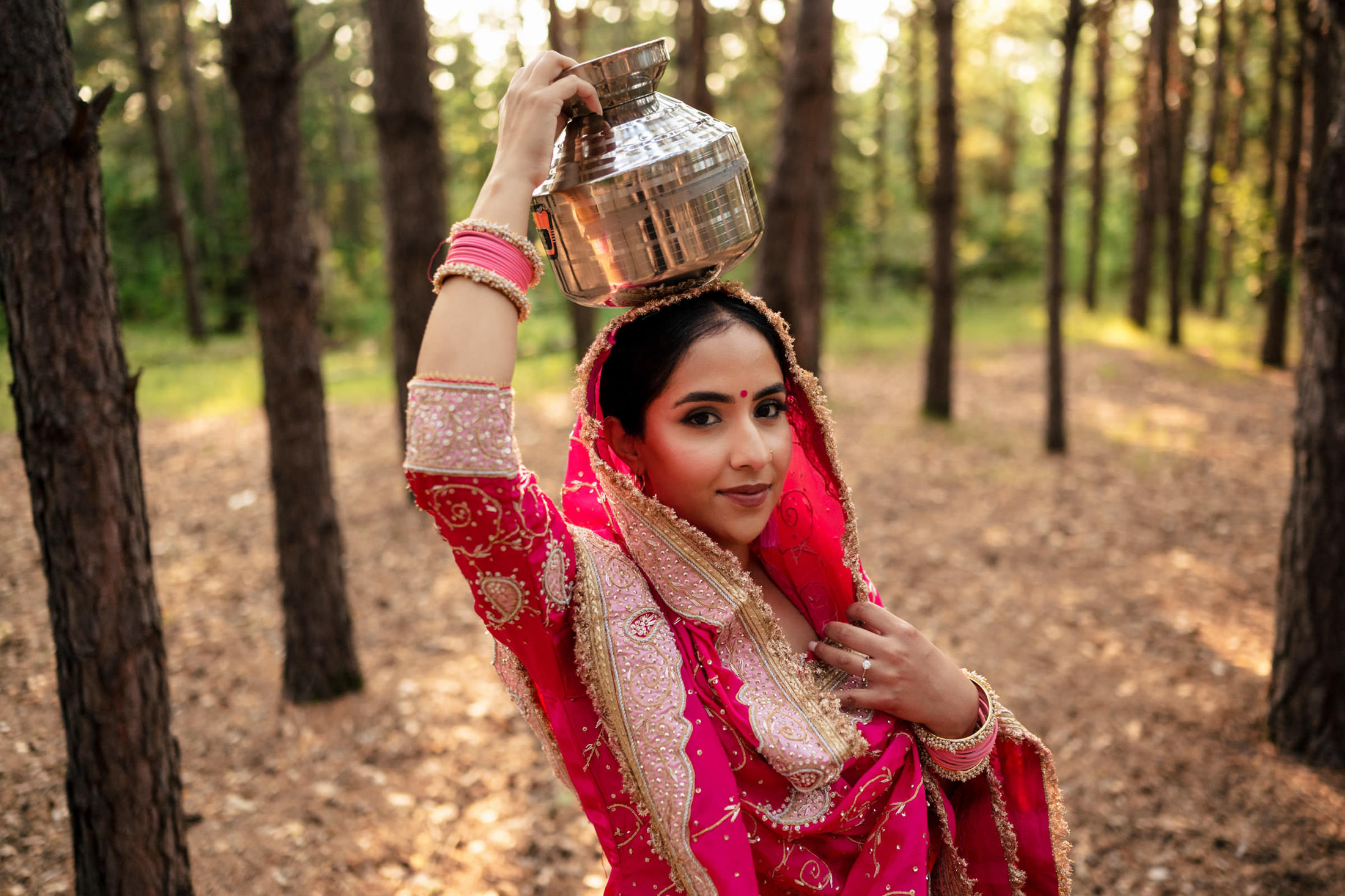 Woman in a pink traditional outfit balances a metal pot on her head in a forest.
