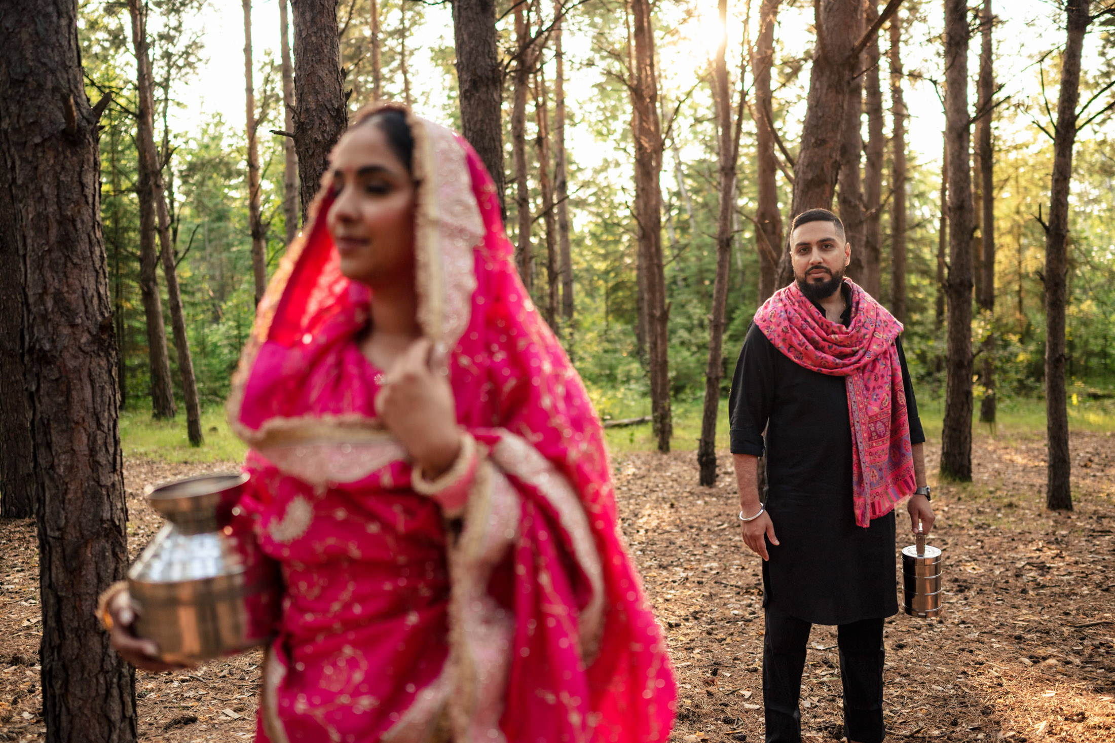 Two people in traditional attire carrying pots walk in a sunlit forest.