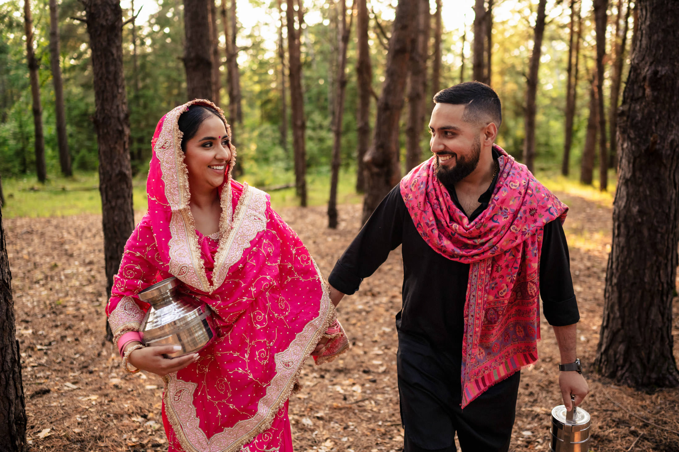 Couple in vibrant attire, carrying pots, walking hand in hand through a sunlit forest.