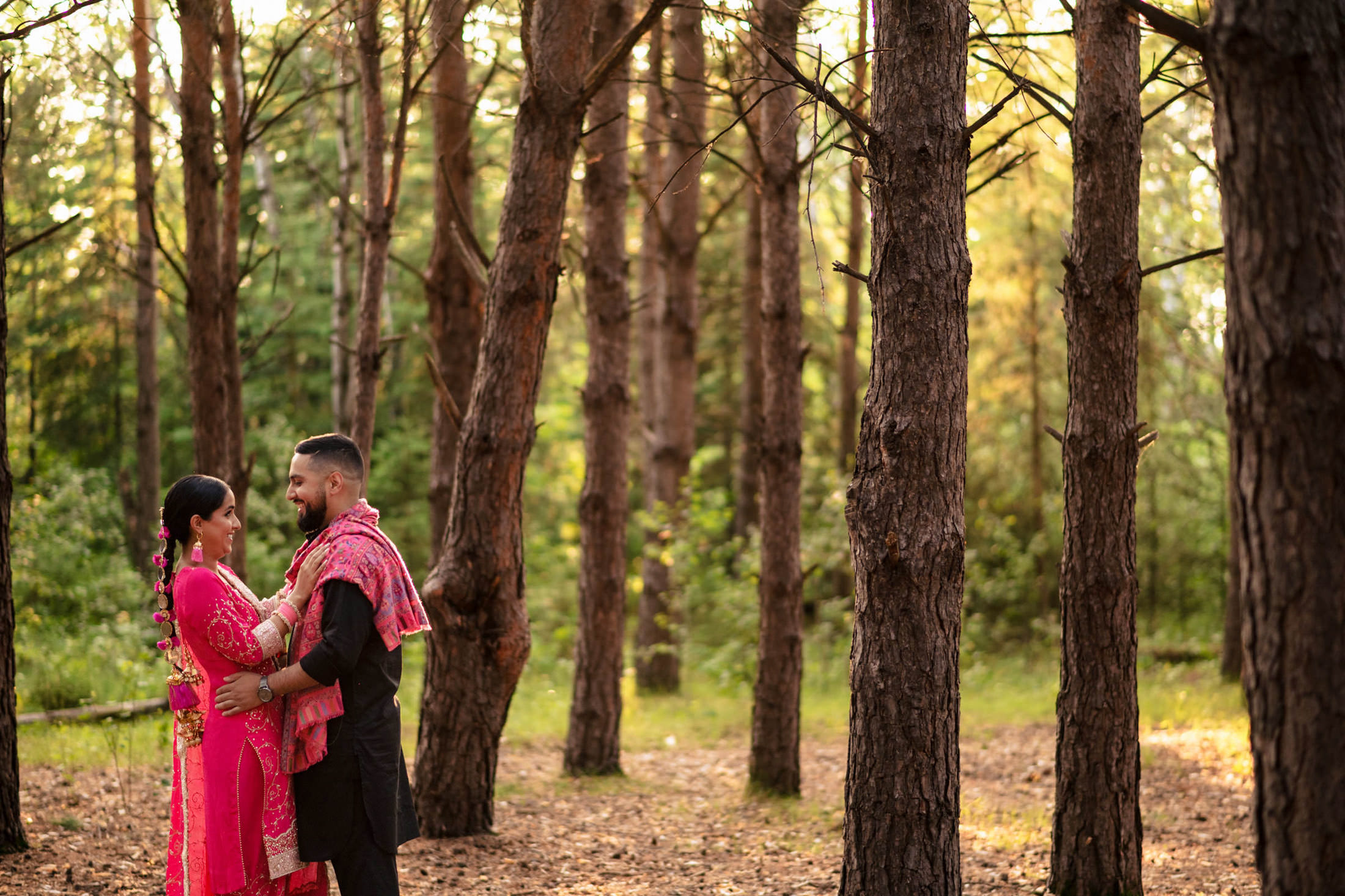 Couple in traditional attire smiling at each other in a sunlit forest.