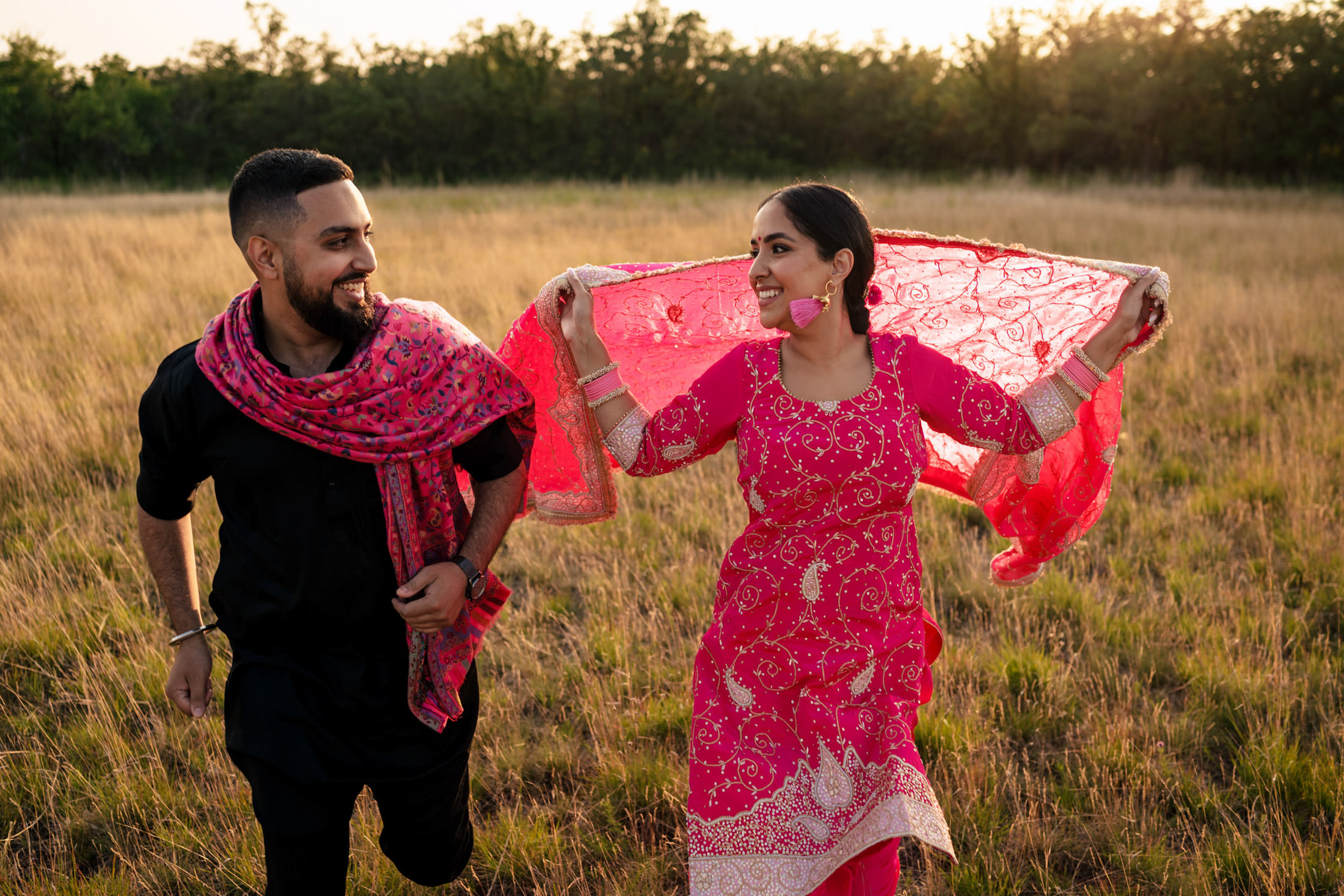 A happy couple in traditional attire joyfully running in a grassy field.