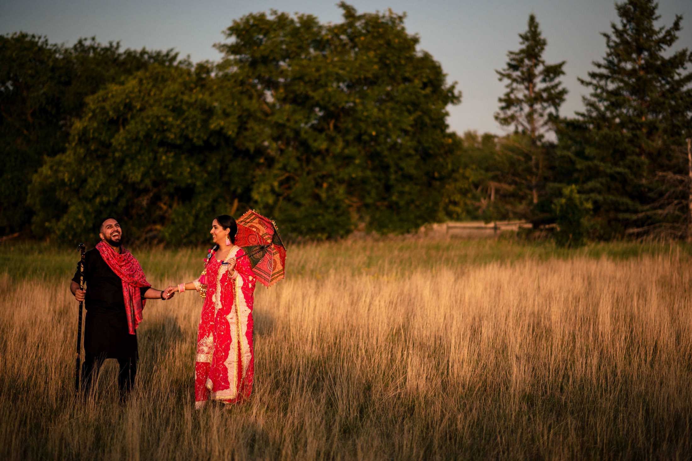 A couple in traditional attire smiles in a grassy field at sunset, holding an umbrella.