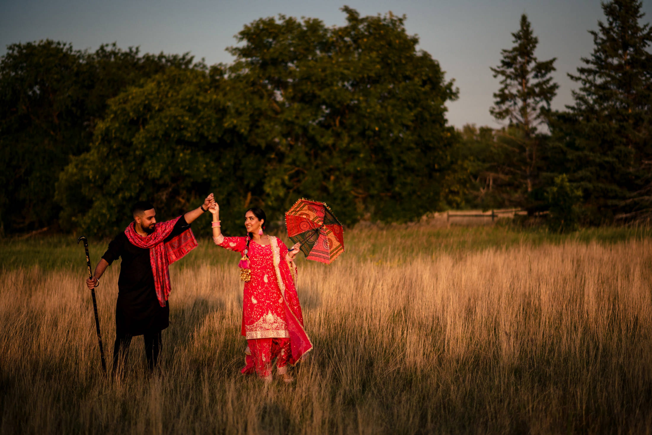 A couple in traditional attire dances in a field, holding an umbrella and a cane.