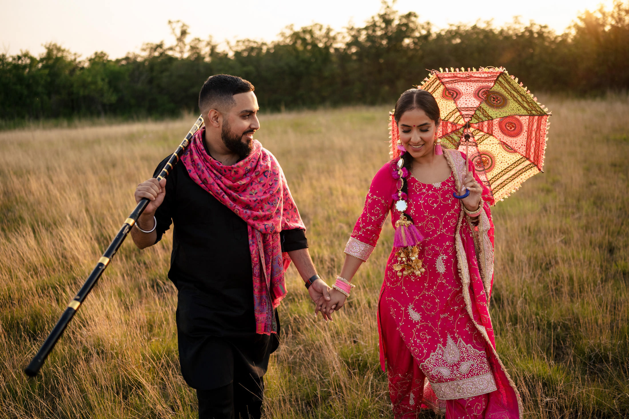 Couple in traditional attire walking in a field, holding hands and an ornate umbrella.