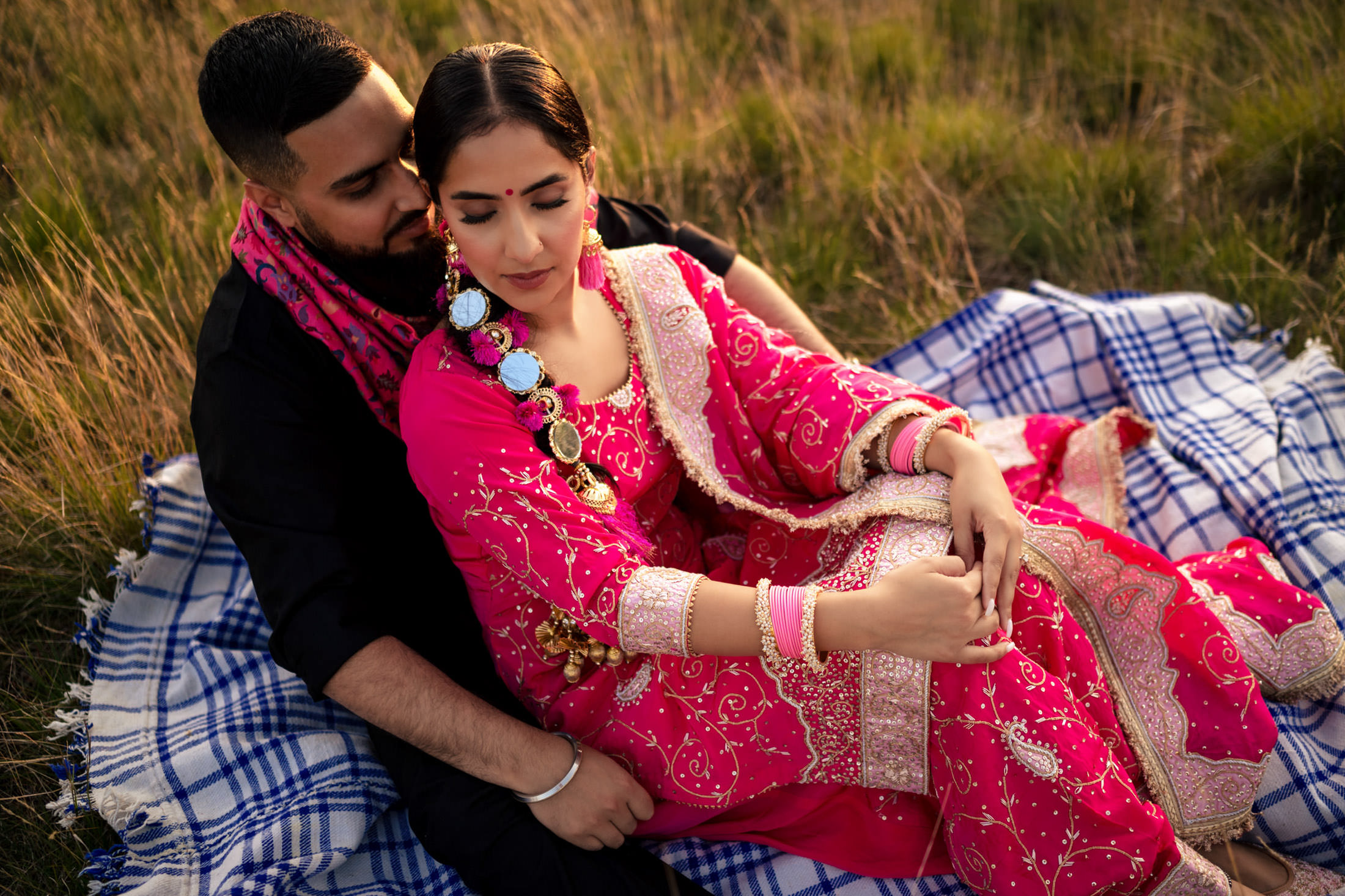 Couple in vibrant traditional attire seated on a blanket in a grassy field.