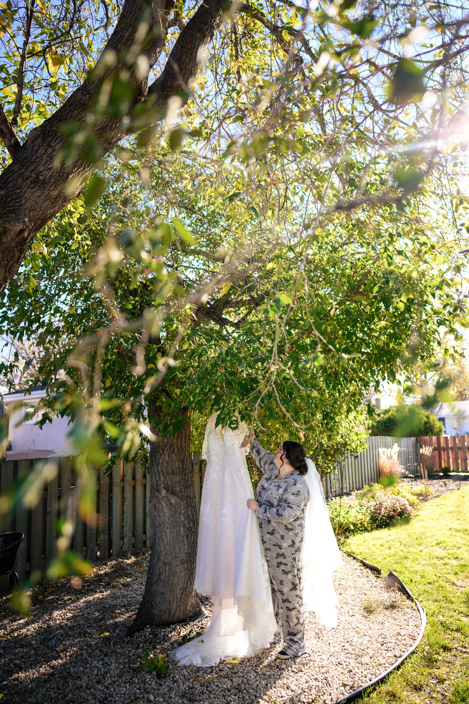 Bride in pajamas admires a wedding dress hanging from a tree in a sunny backyard.