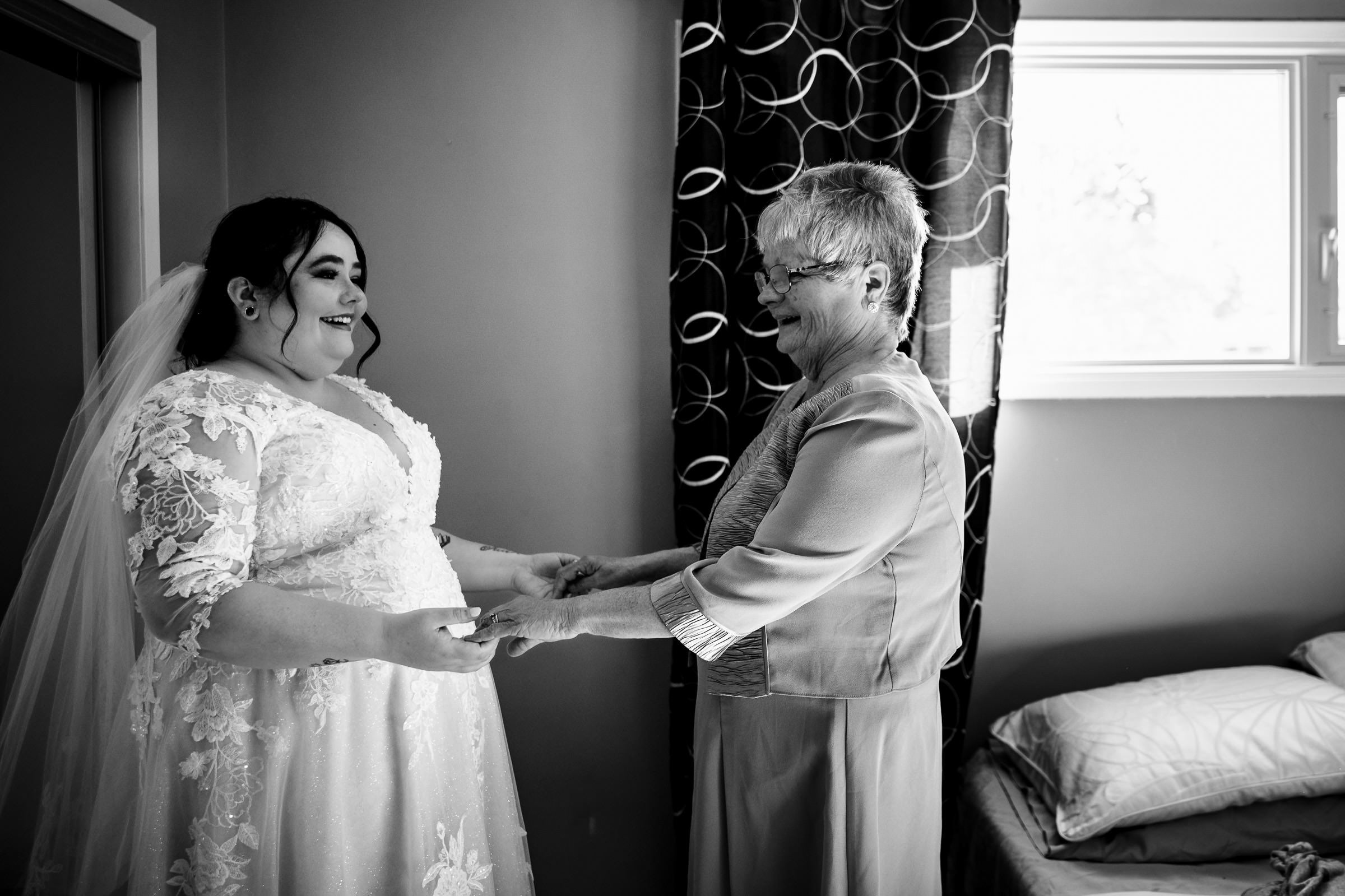 Bride and older woman smile and hold hands in a room.