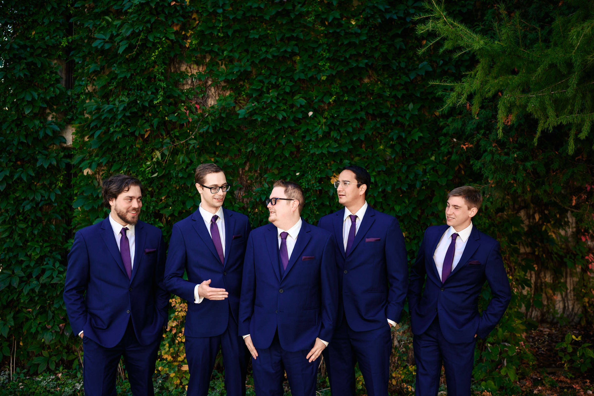 Five men in blue suits stand in front of a leafy wall, engaged in conversation.