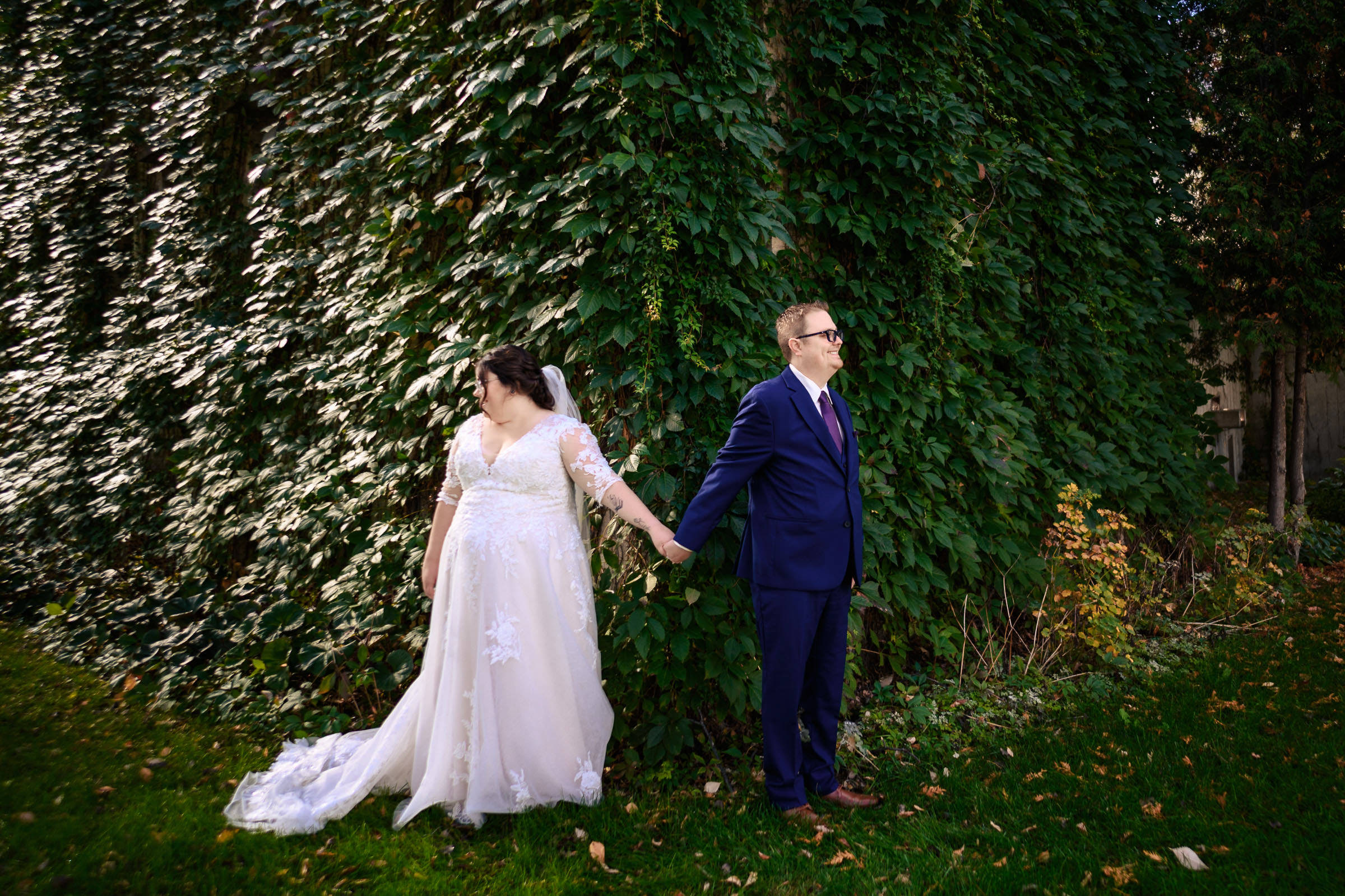A bride and groom hold hands, standing back-to-back near a wall covered with greenery.