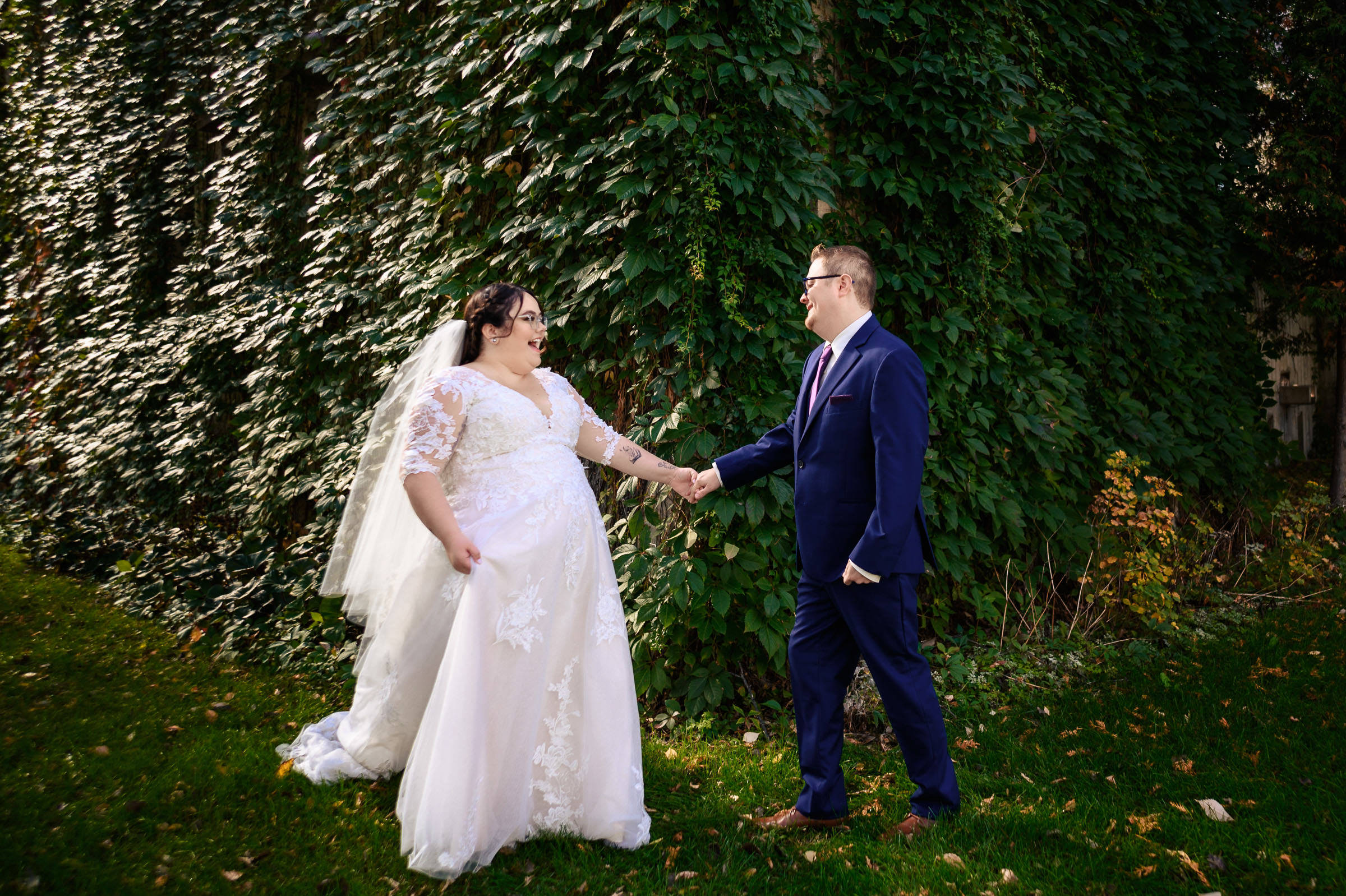 Bride and groom holding hands in front of lush green ivy wall.