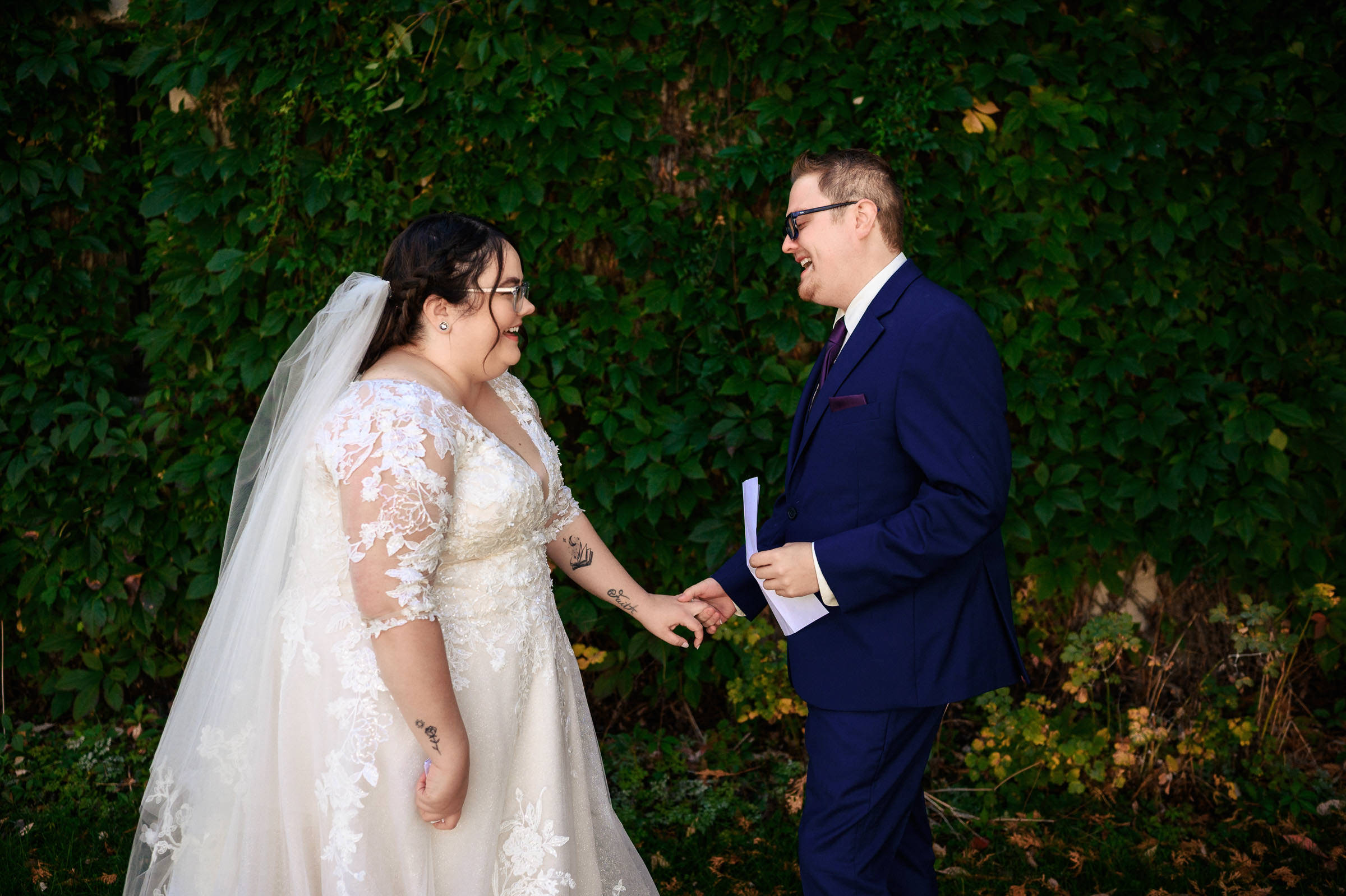 Bride and groom smile at each other in front of lush green foliage.