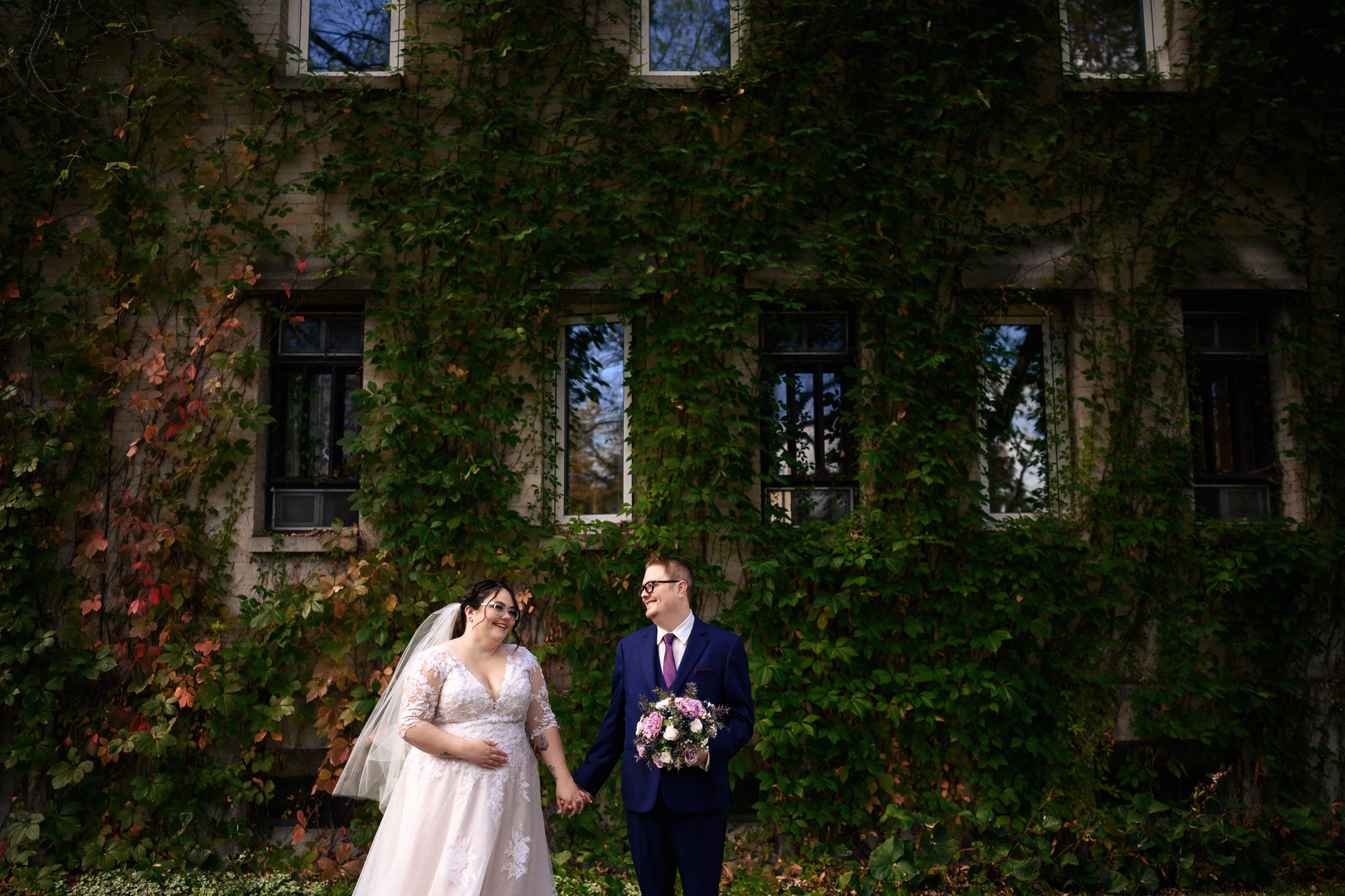 Bride and groom hold hands in front of a vine-covered building.