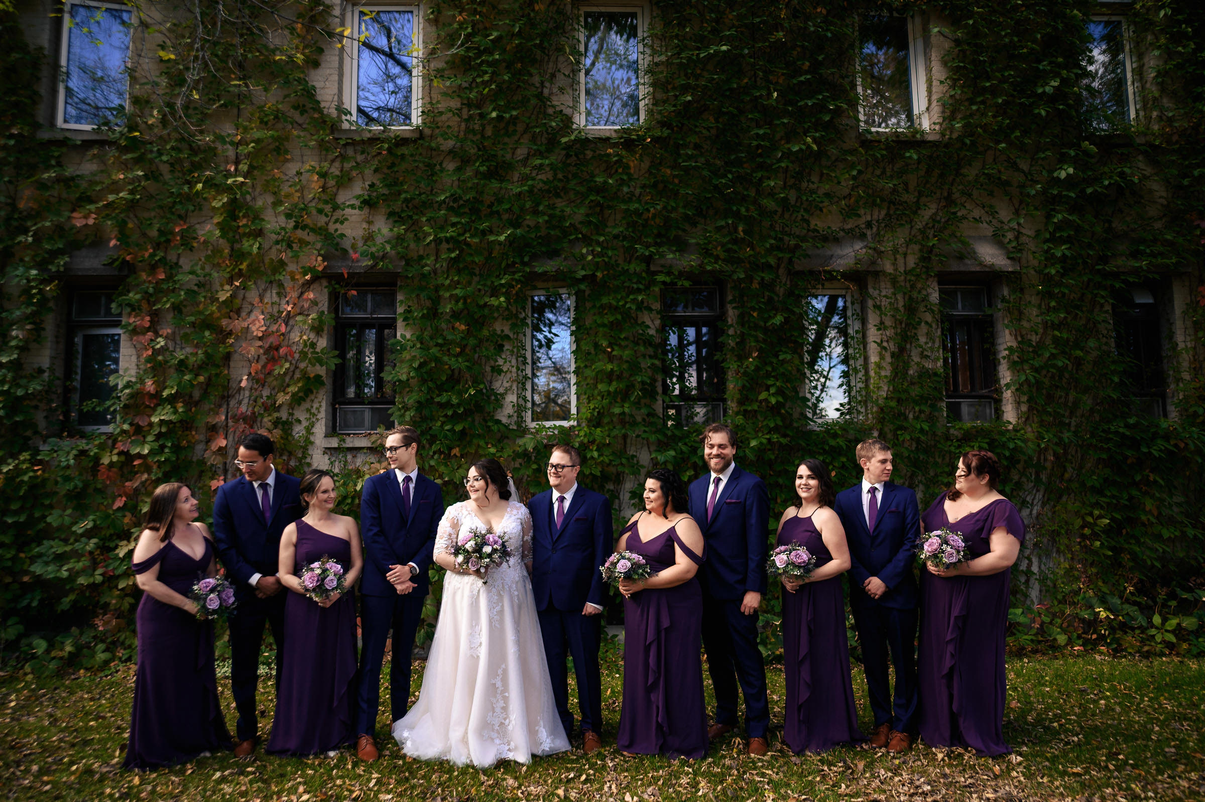 Bride, groom, and wedding party pose outdoors in front of an ivy-covered building.