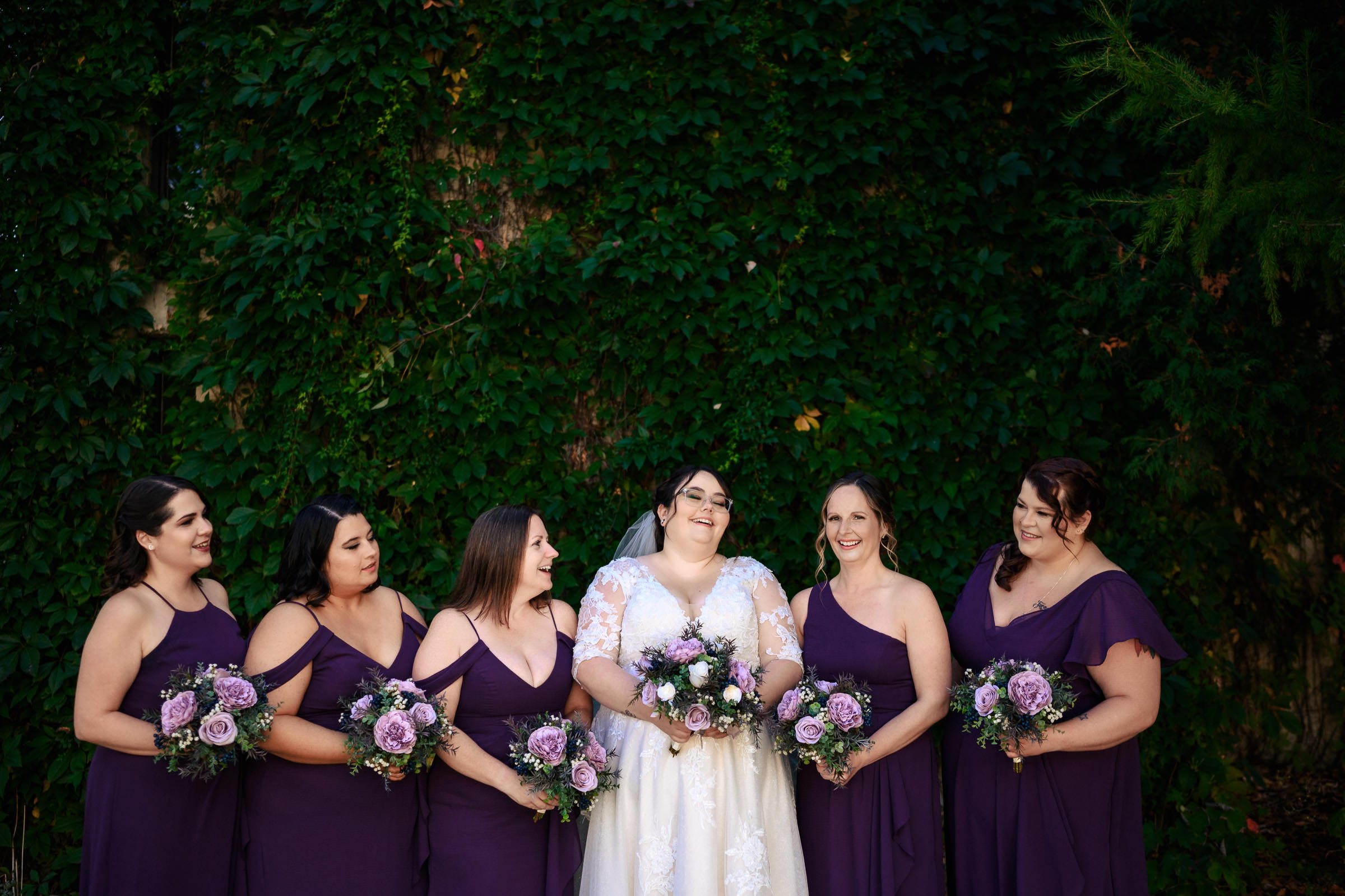Bride with five bridesmaids in purple dresses holding bouquets in front of greenery.