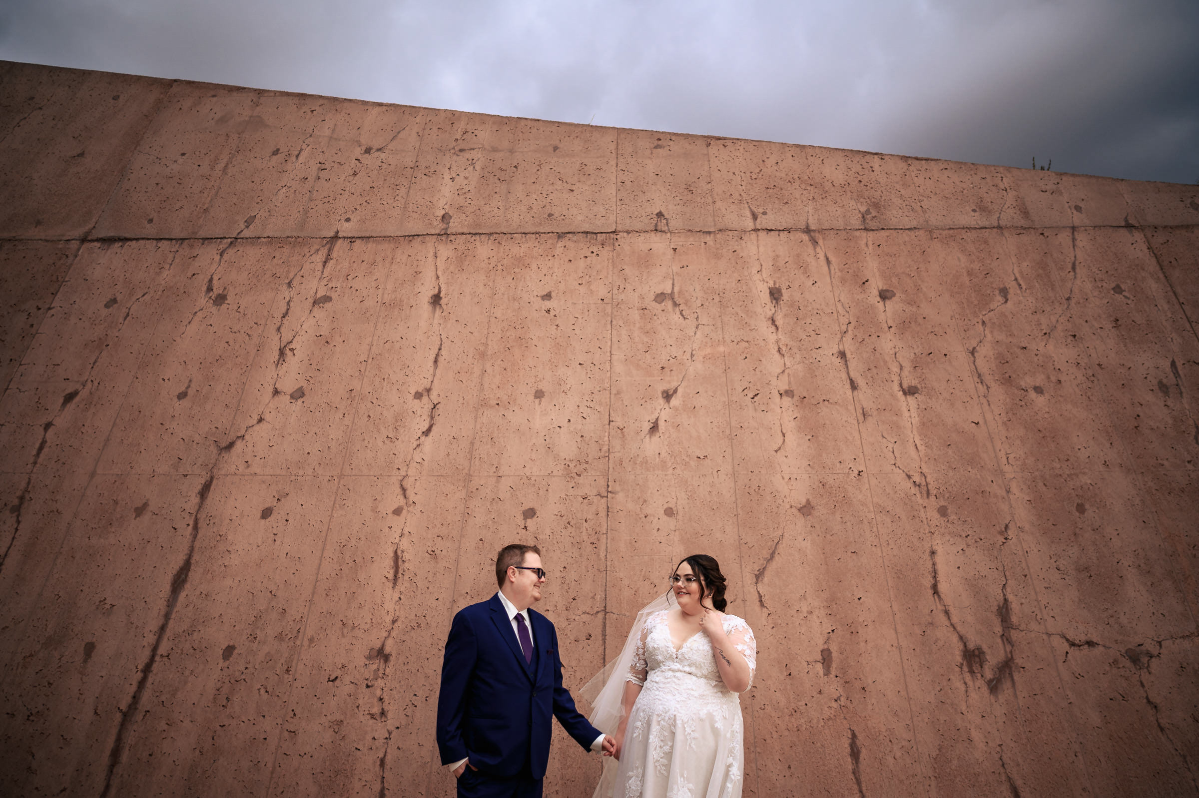Bride and groom standing in front of a large textured wall, holding hands.