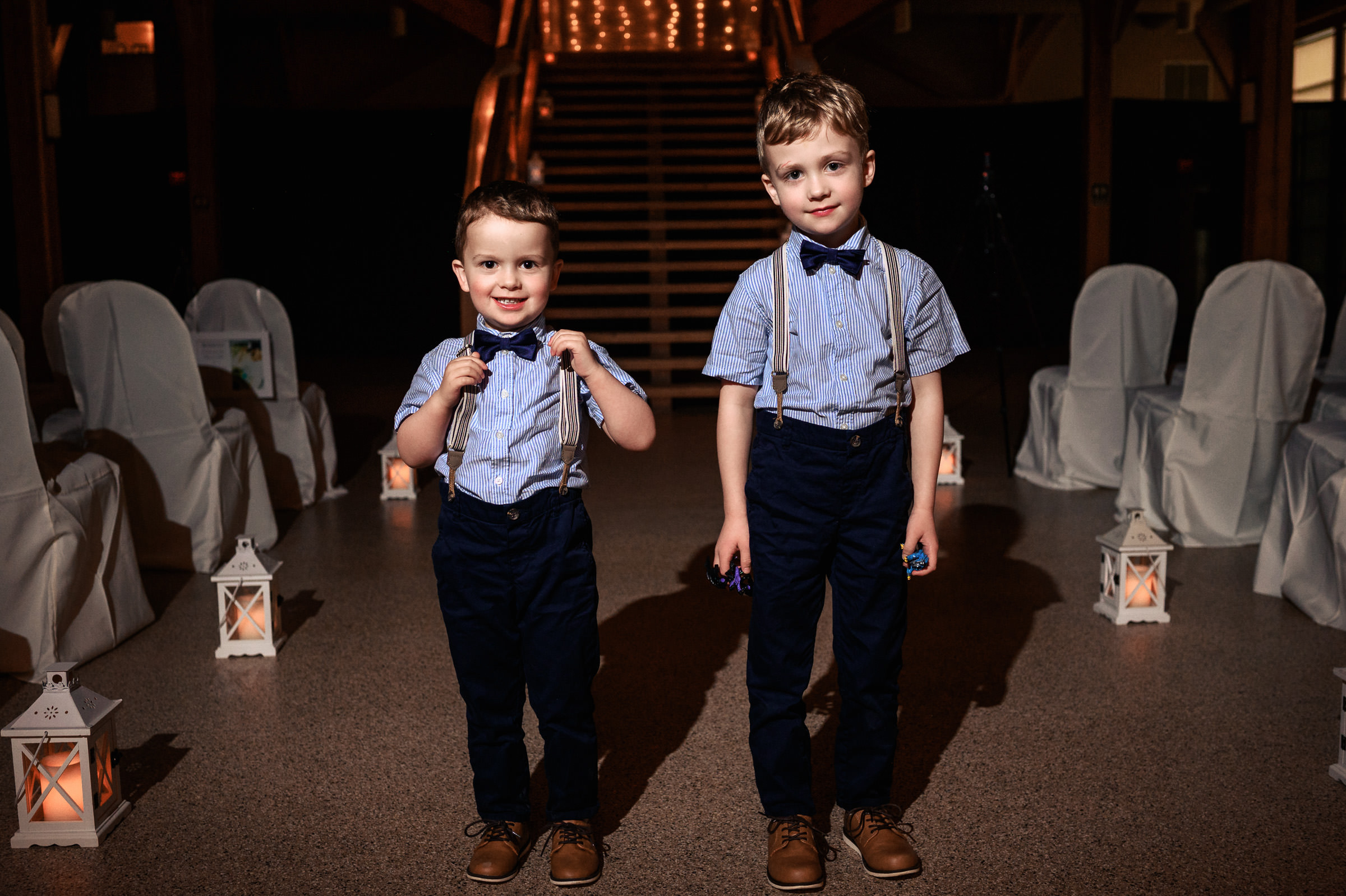 Two young boys in matching outfits stand indoors, each wearing a bow tie and suspenders.