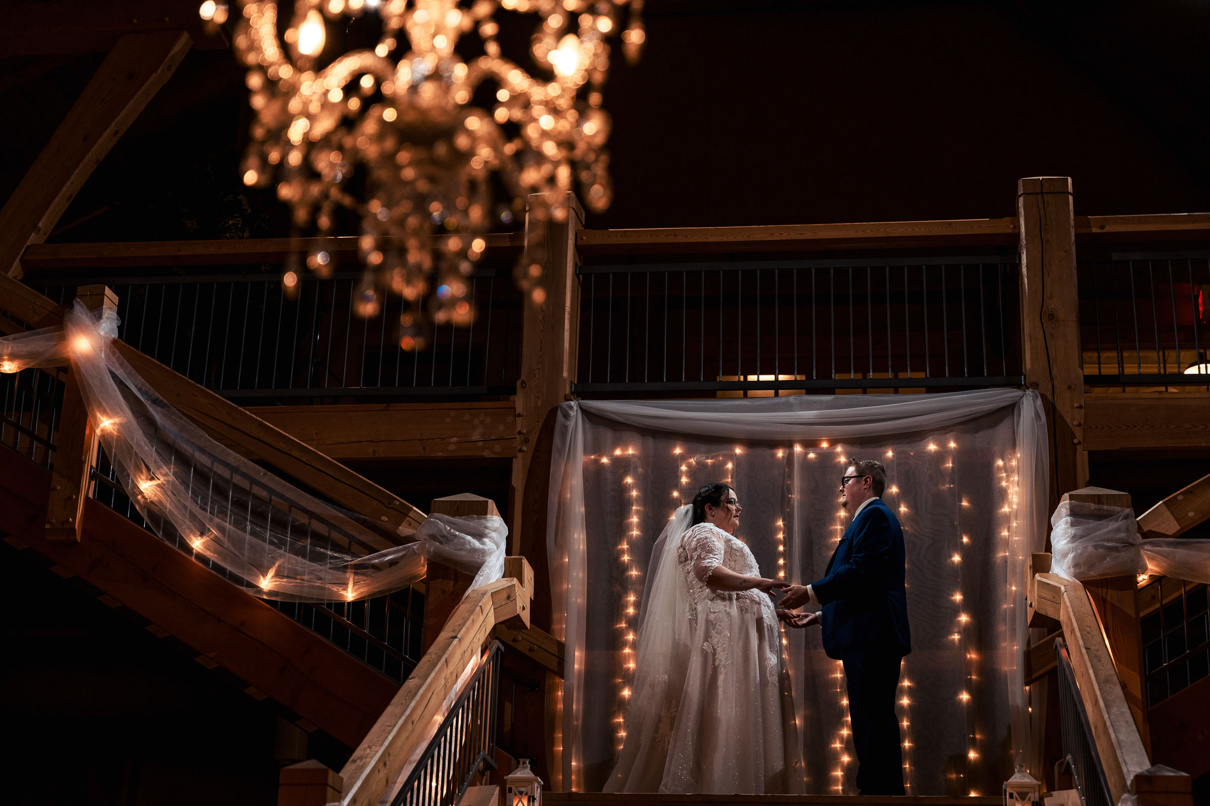 Couple holds hands under string lights on a decorated staircase with a chandelier above.