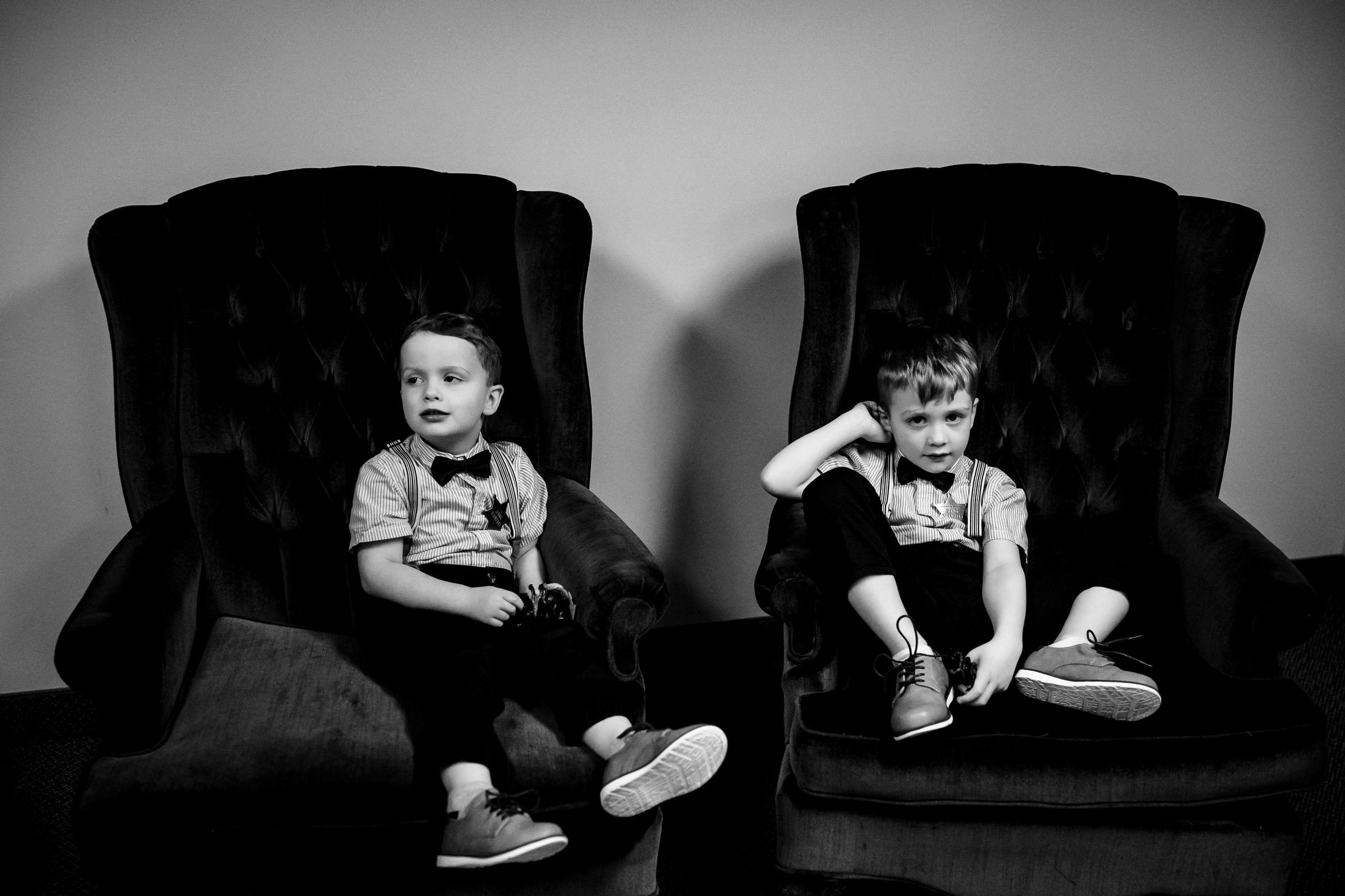 Two young boys sit in oversized chairs, dressed in matching attire, with neutral expressions.